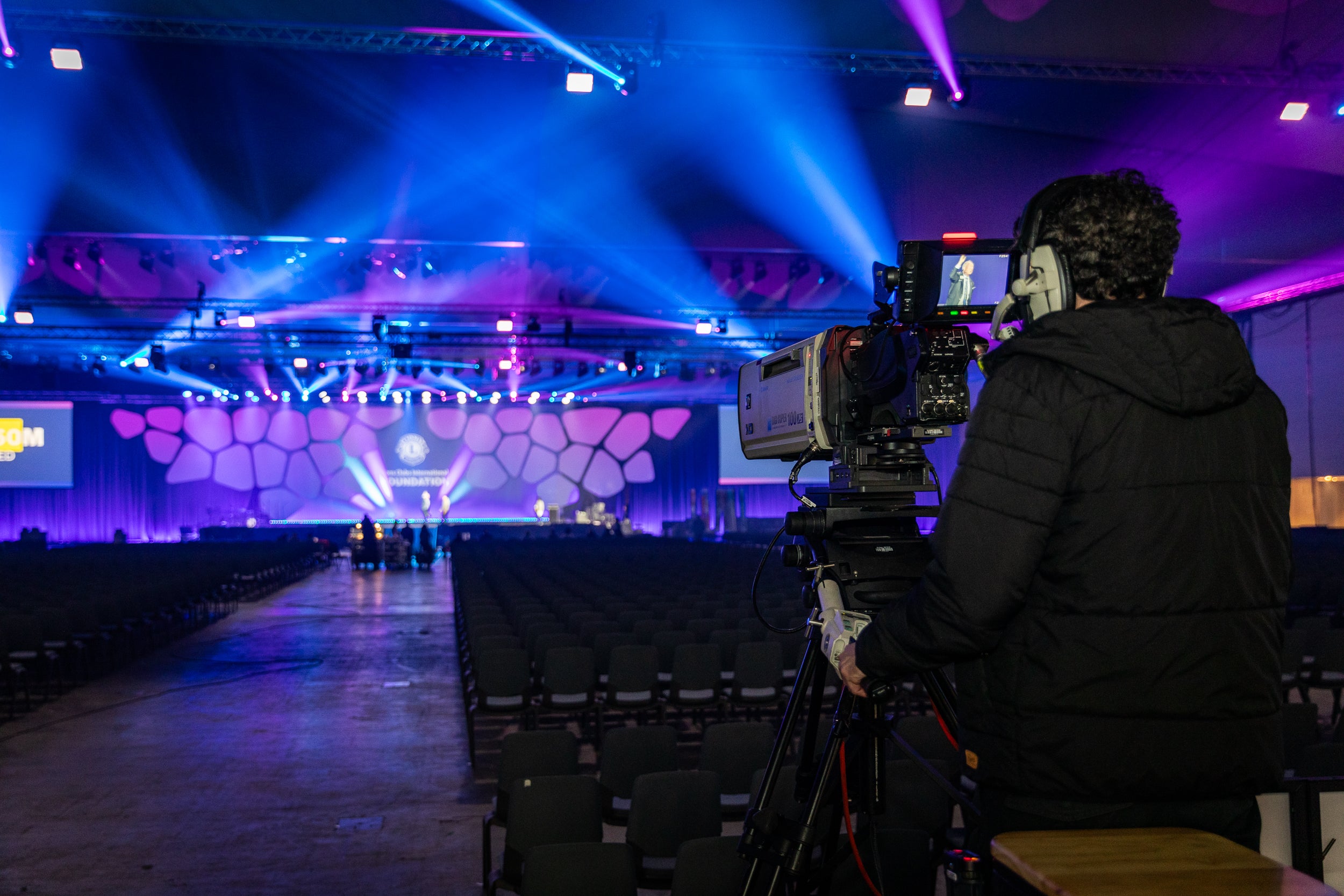 Person operating a camera in a large auditorium with stage and audience seating.