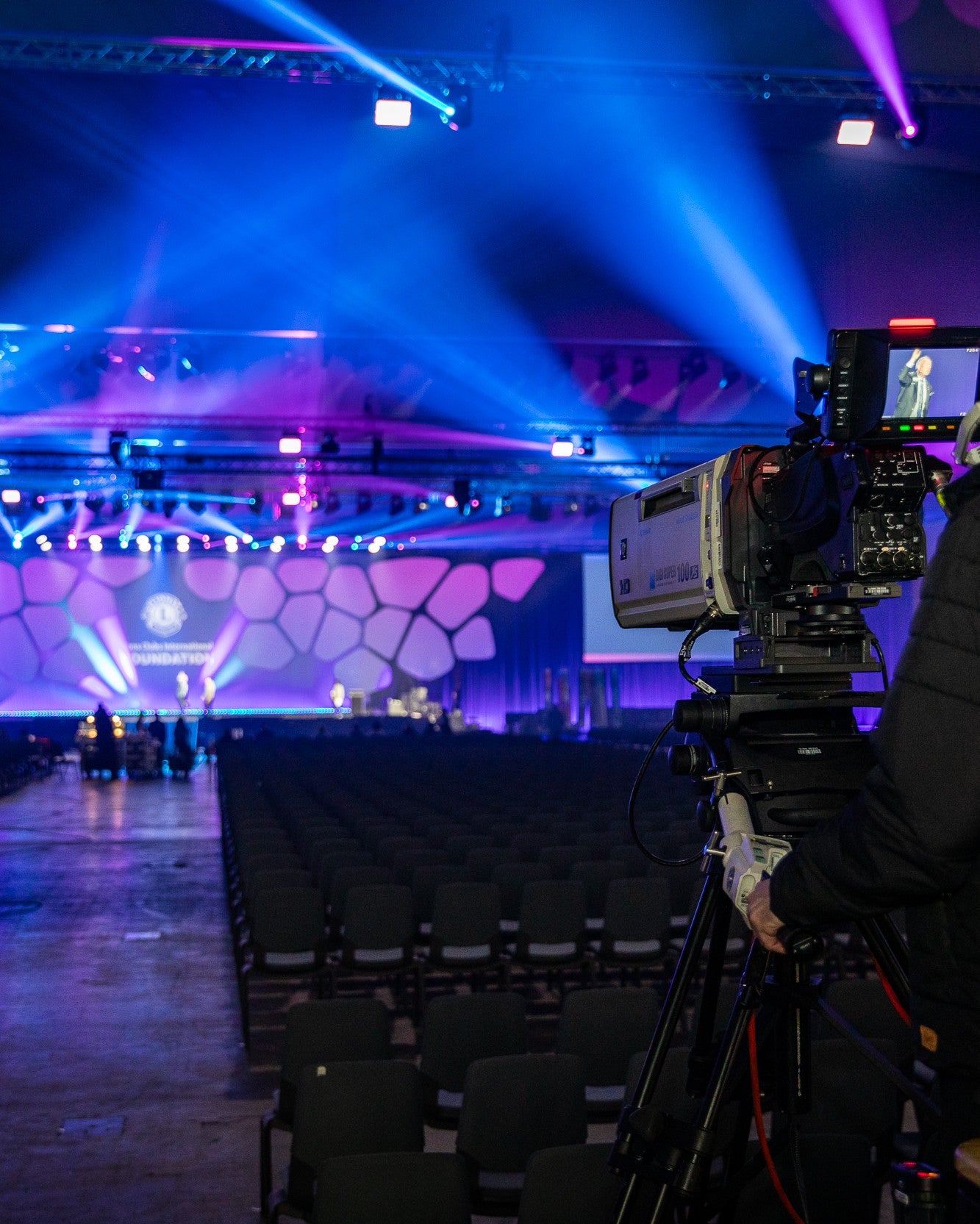 Camera on a tripod facing an auditorium with stage lights and a screen.