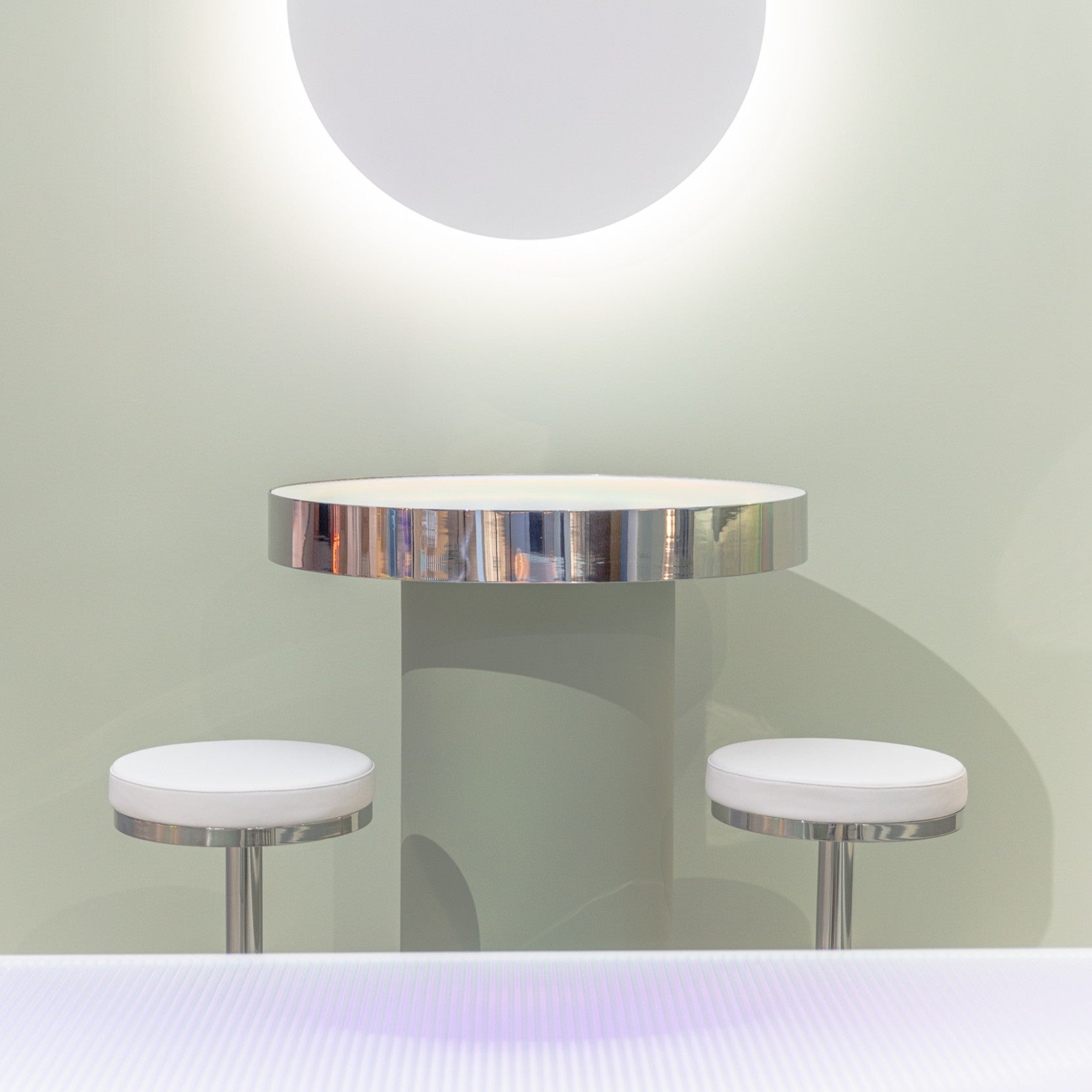 Zeon round table with chrome rim and two stools against a light green wall.