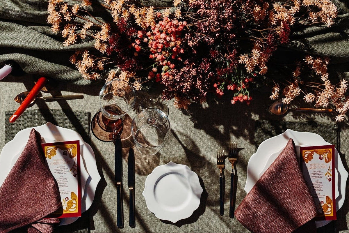 An overhead view of a styled dining table setting with white plates, dark metal cutlery, folded burgundy napkins, pink taper candles, and a central dried floral arrangement on a textured tablecloth.