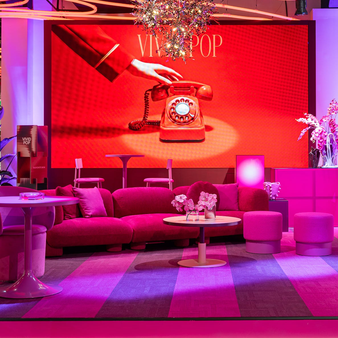 A vibrant pink and red lounge room set with a striped rug, pink velvet sofas, matching armchairs, and a large red backlit screen displaying a retro rotary phone graphic.