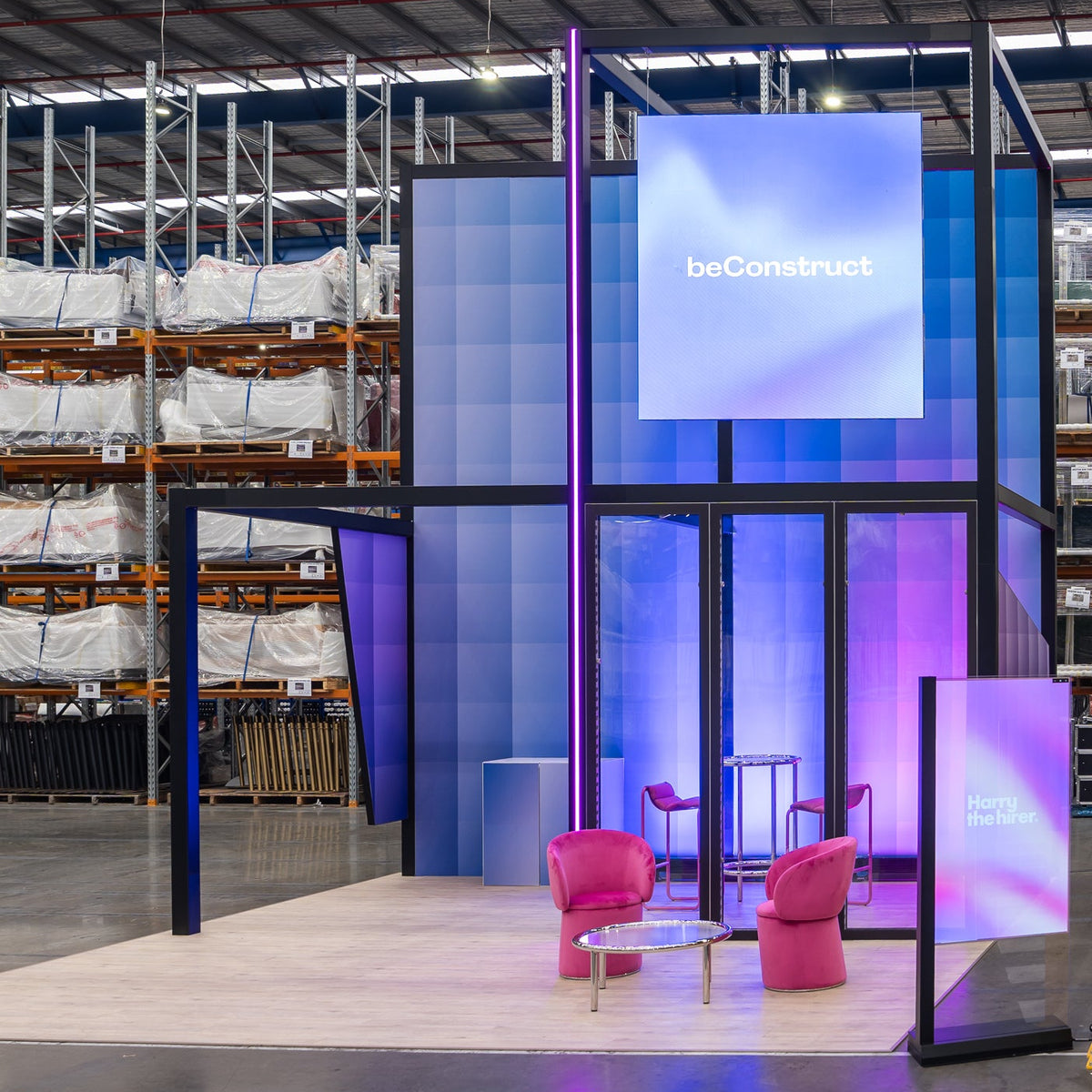 A modular event pavilion or exhibition booth with a blue and purple illuminated frame, pink chairs, and a large screen displaying 'beConstruct', set up inside a large warehouse with shelving units in the background.