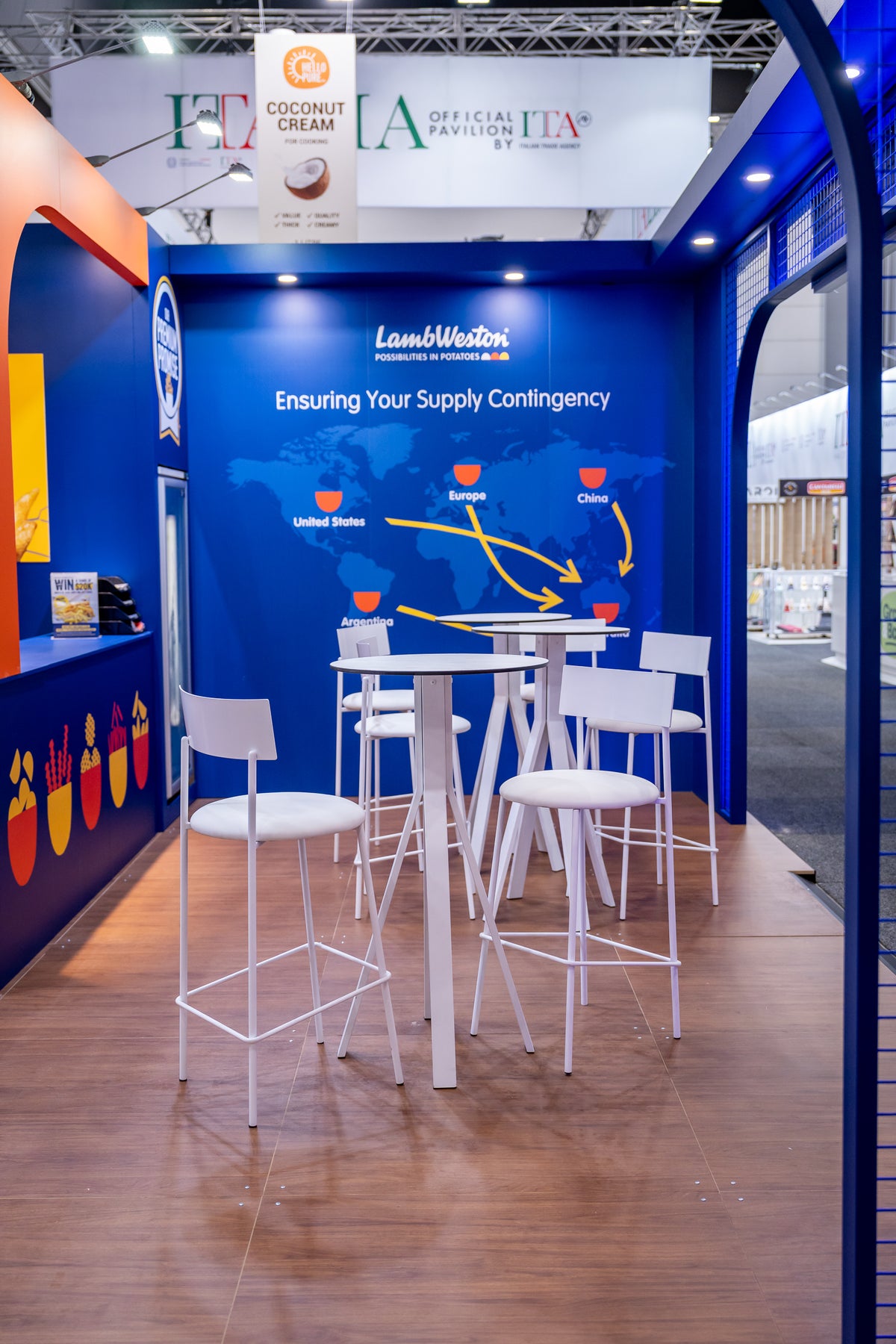A modern booth interior with a blue branded wall, white high-top tables, and white bar stools. Visible branding includes Lamb Weston on the back wall and a large ITA sign overhead.