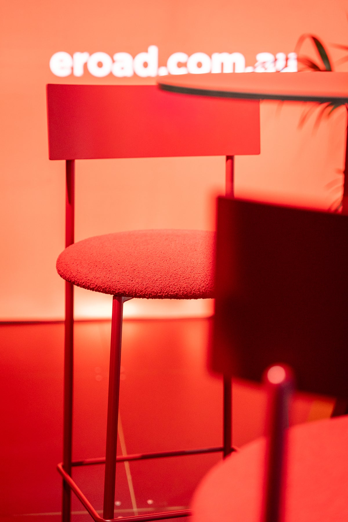 A modern red upholstered bar stool with a slim metal frame, shown in a dimly lit setting with a red-orange ambient glow. A white 'eroad.