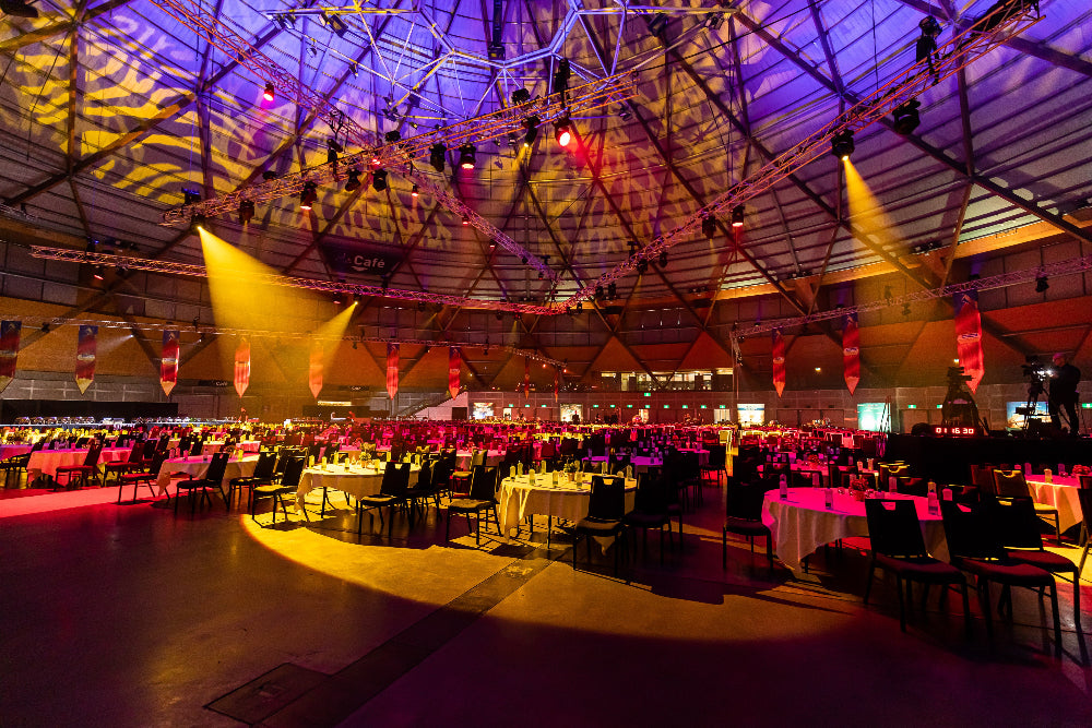 Large indoor event space with tables and chairs set up for an audience, illuminated by colorful lights.