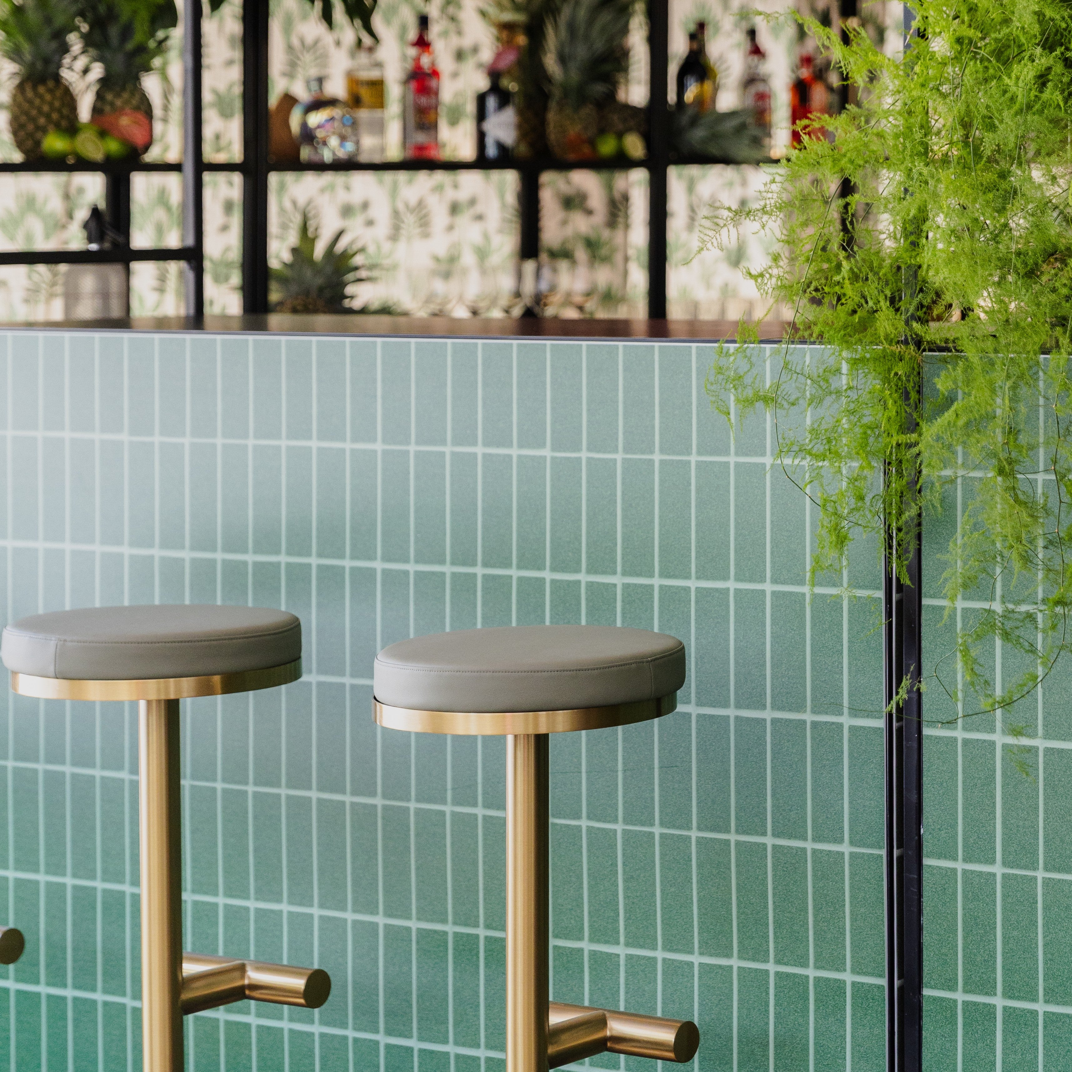 Two bar stools with gold legs and green cushions in front of a green tiled wall.