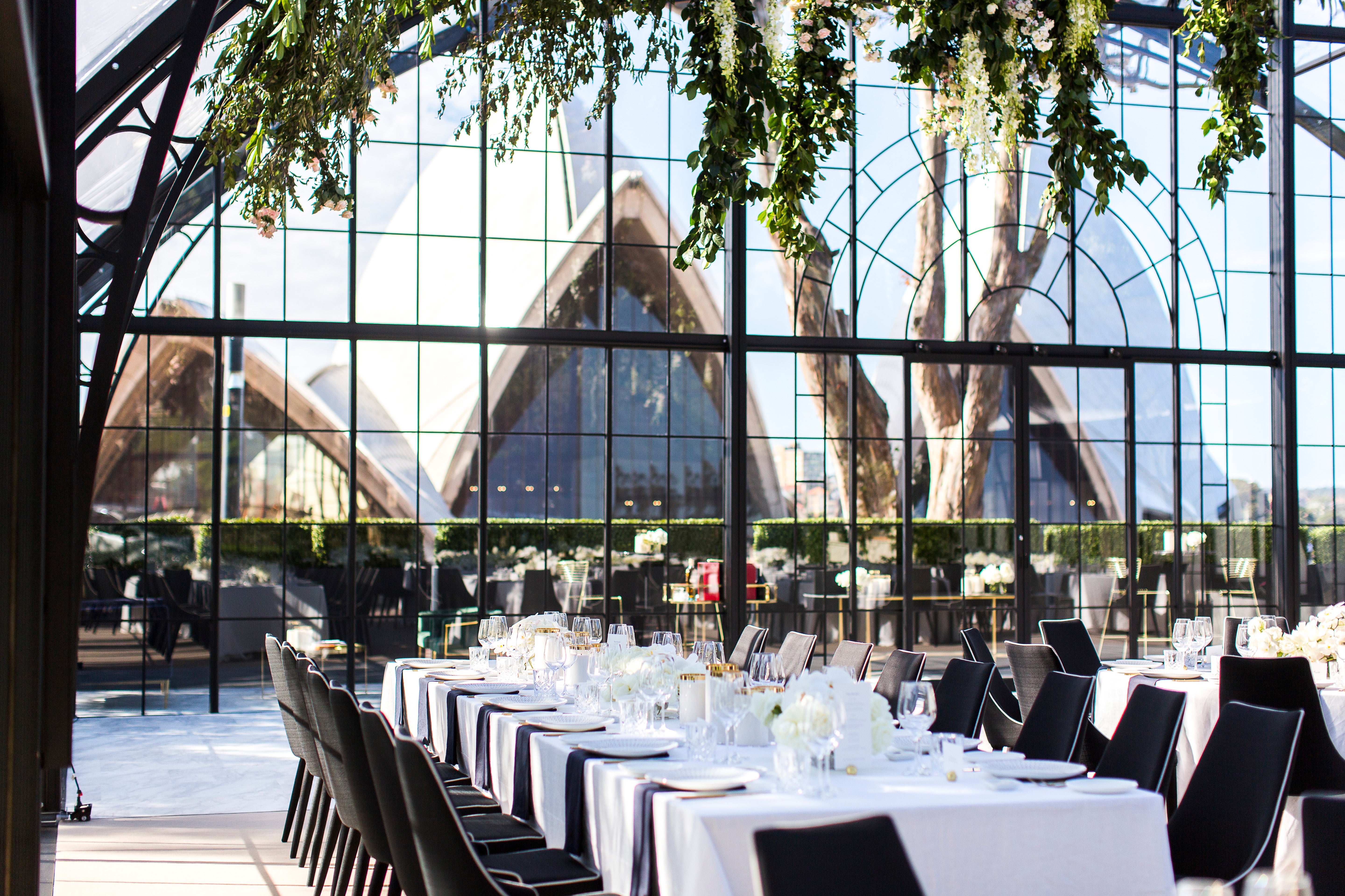 Dining setup with tables and chairs in front of a large glass wall with the Sydney Opera House in the background. Setup is for a wedding.