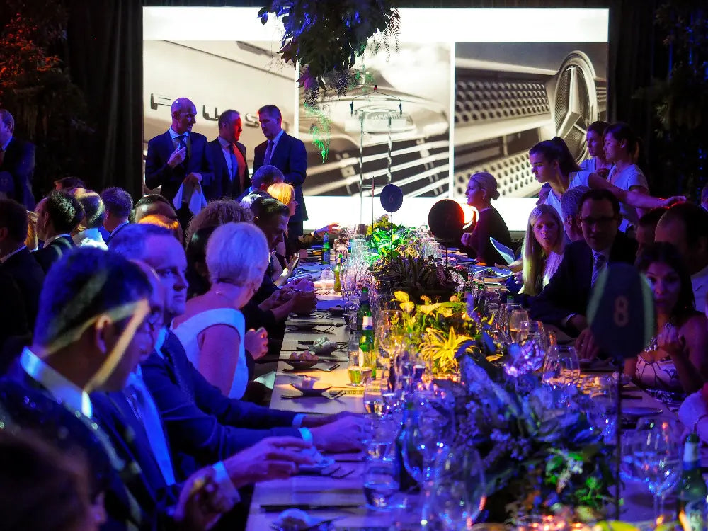 Dinner event with guests seated at a long table, featuring Mercedes-Benz vehicles in the background.