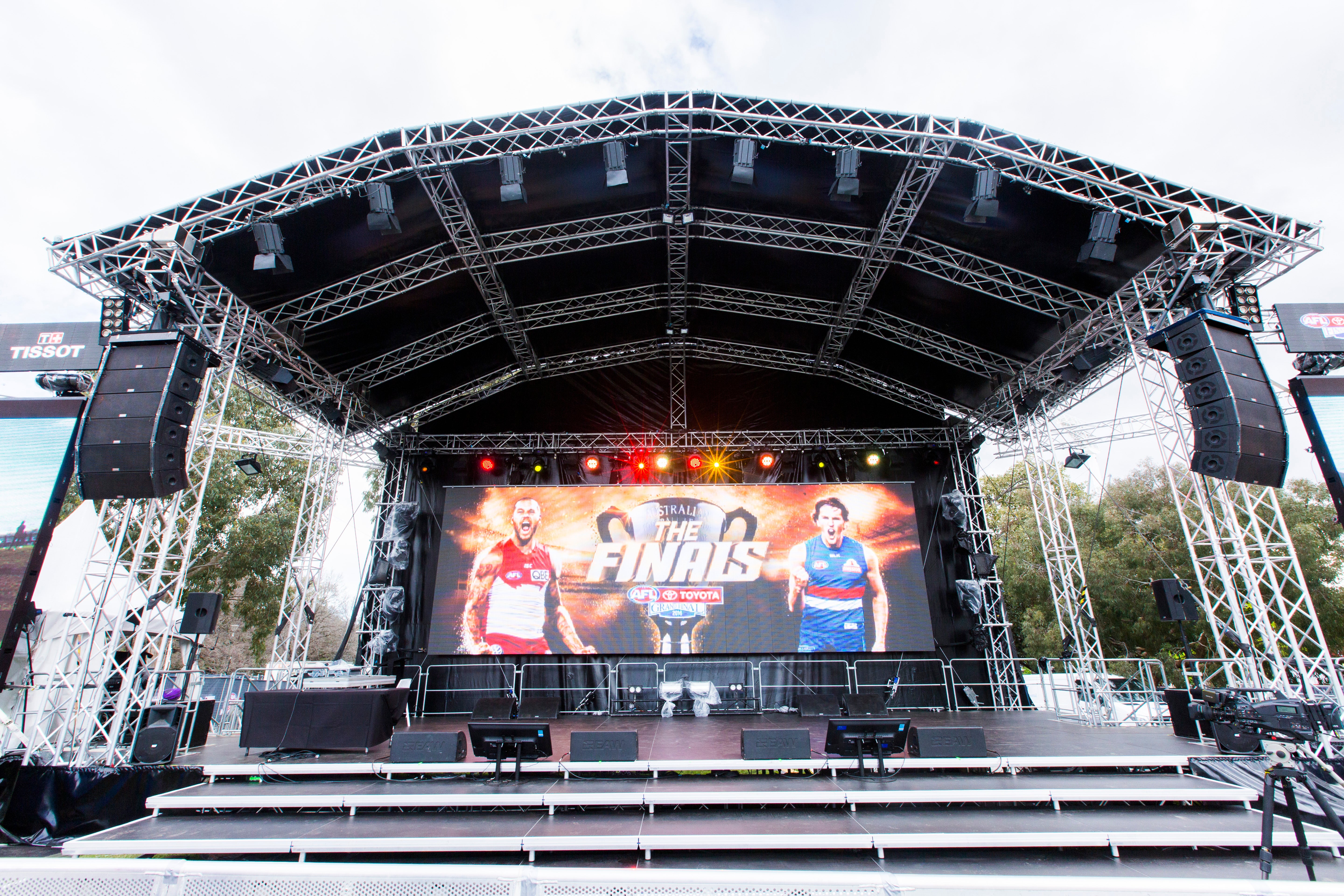 Outdoor stage setup with a large screen displaying two AFL athletes, surrounded by trees and clear sky.