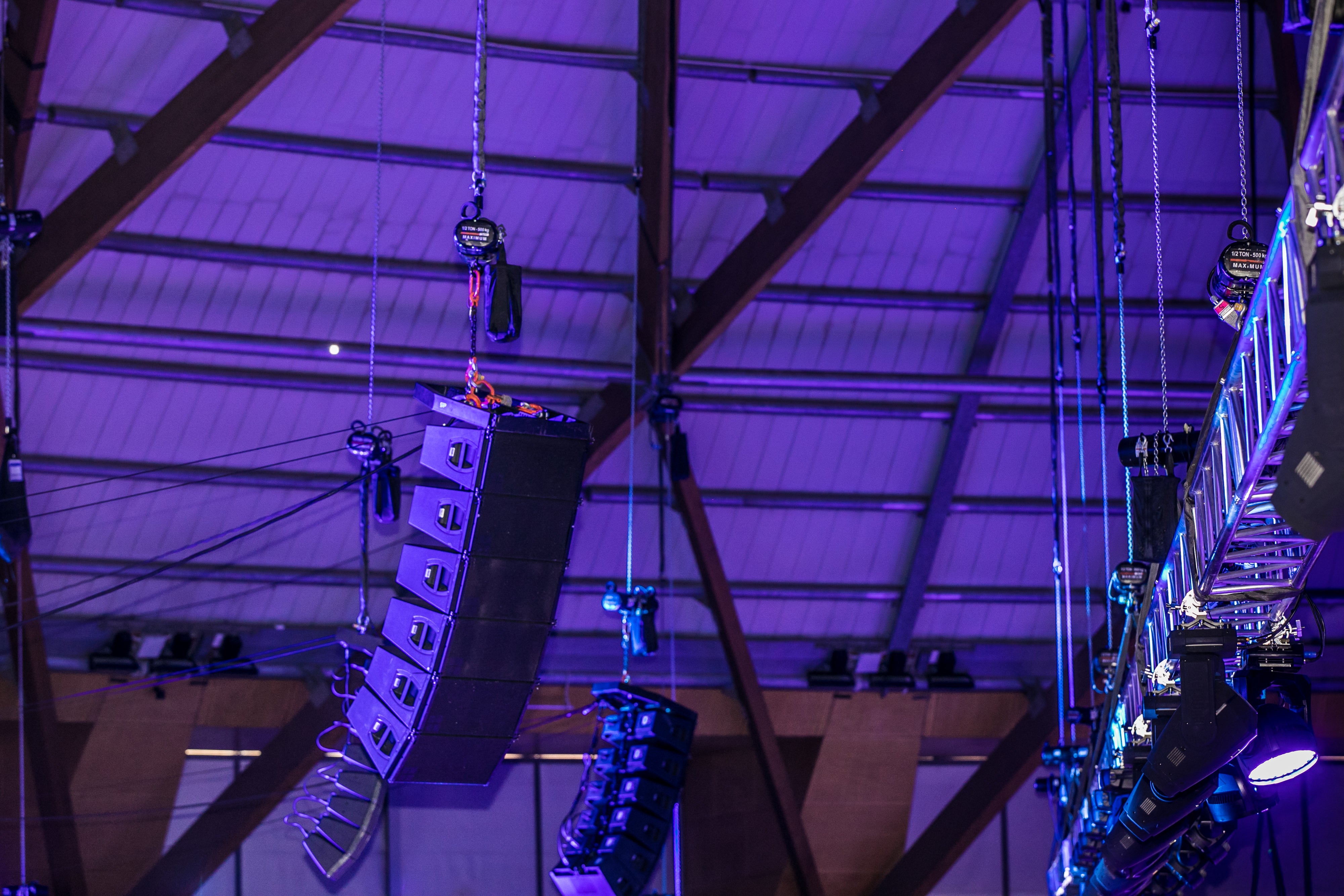 Sound equipment hanging from the ceiling in a large indoor venue with purple lighting.