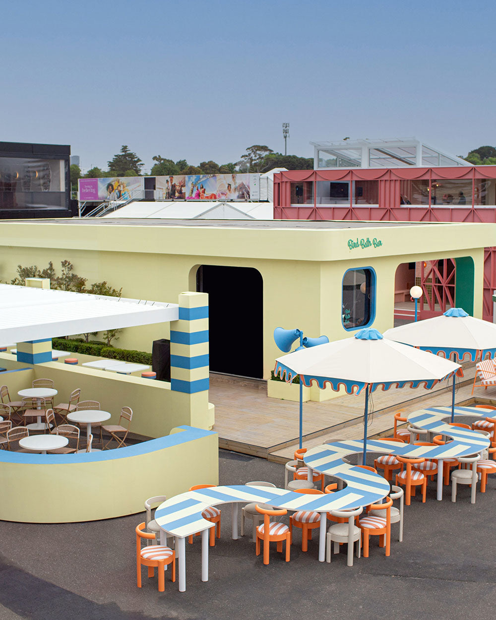 Outdoor seating area with tables, chairs, and striped umbrellas in a colorful custom building setting at Spring Racing Carnival