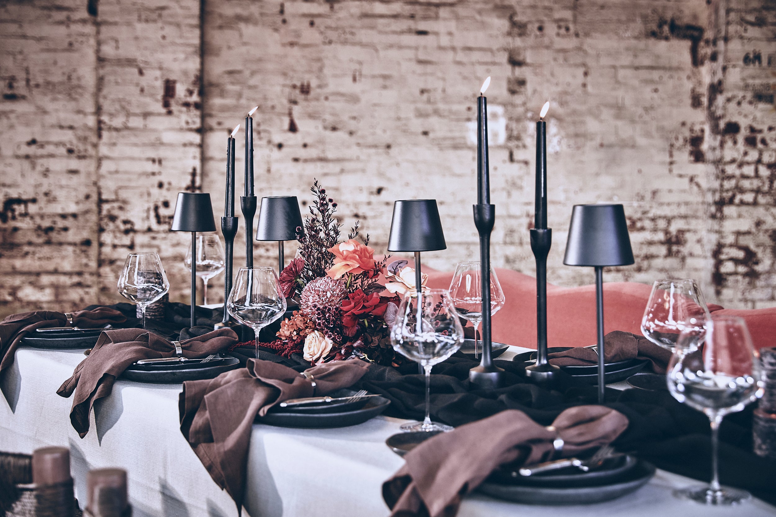 Elegant table setting with black candles, glasses, and a floral centerpiece against a brick wall.