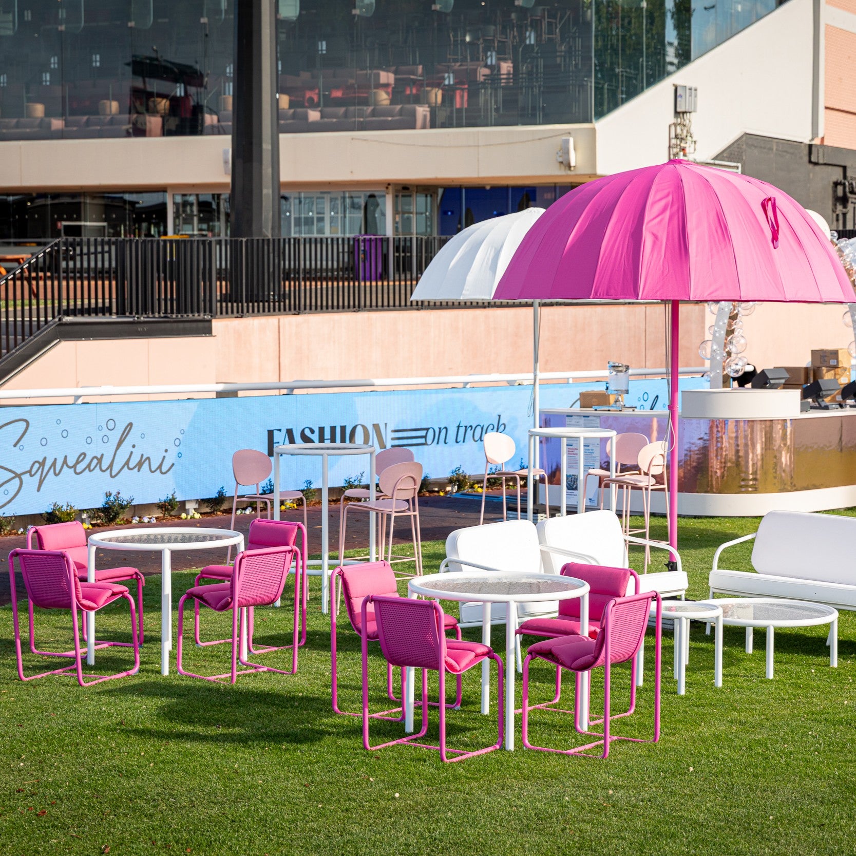 Outdoor seating area with pink chairs and a large pink umbrella on a grassy field, with a building and branding in the background.