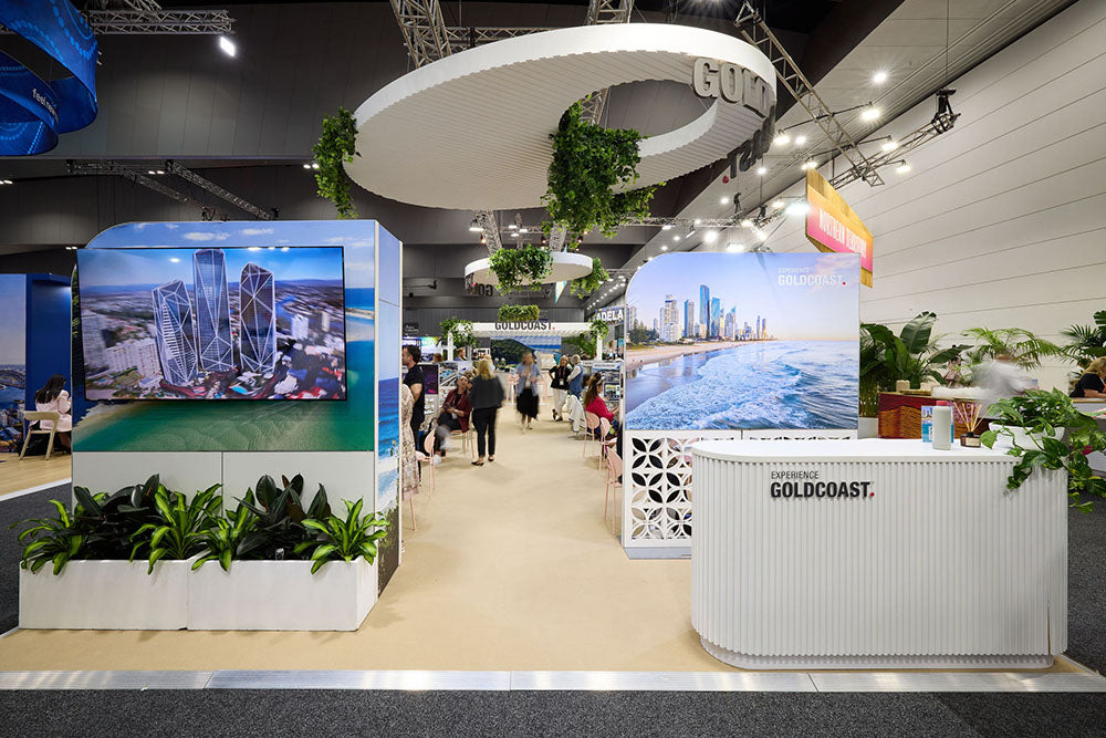 Exhibition stand with large screens displaying cityscapes, surrounded by plants and people walking around.