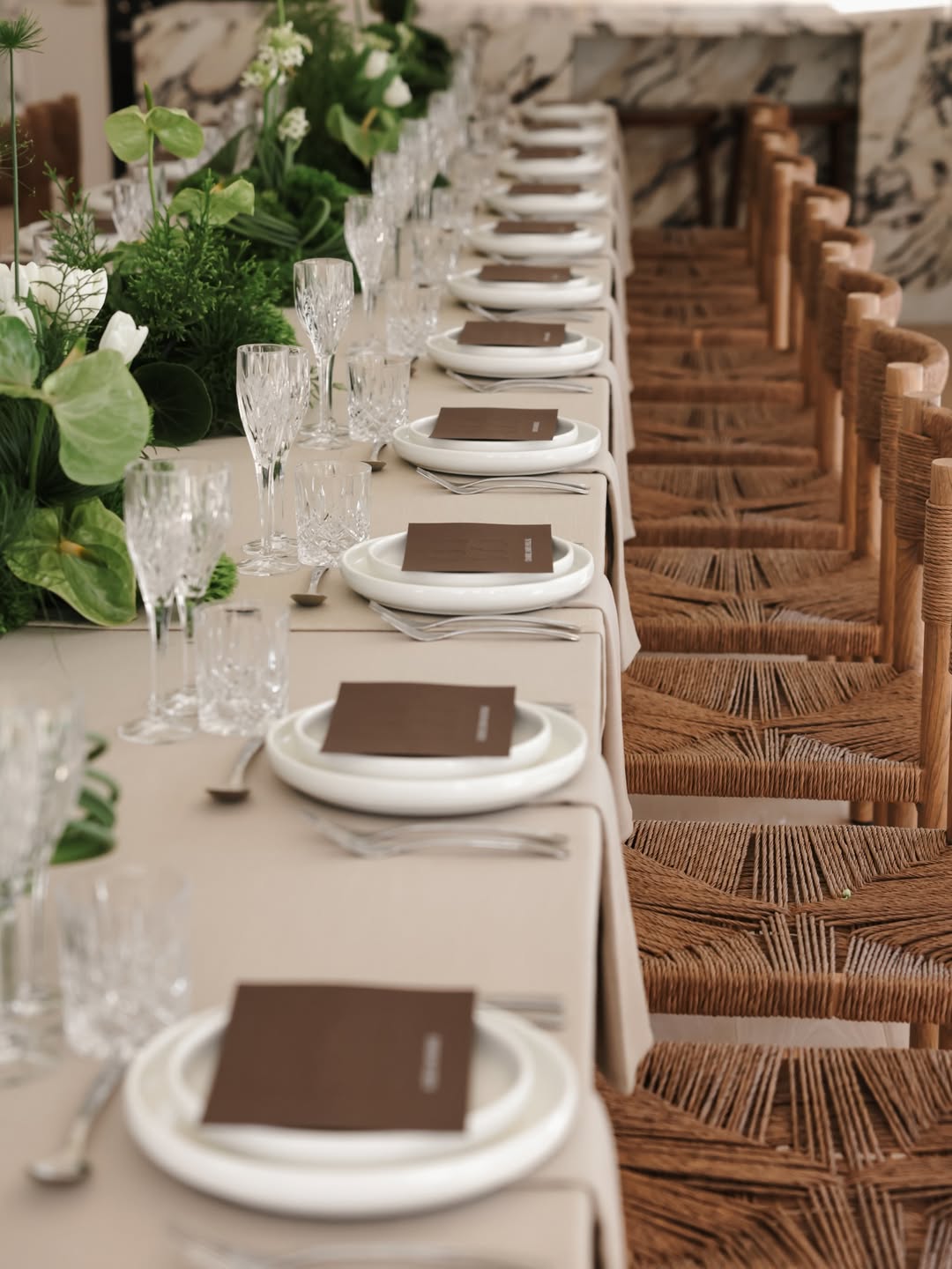 Elegant dining setup with white plates, brown napkins, and clear glasses on a long table with wicker chairs.