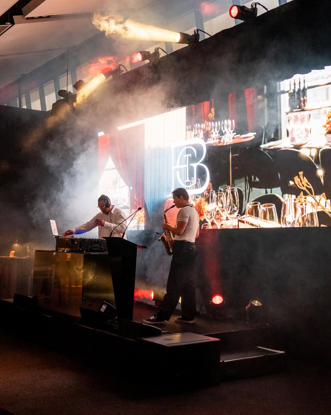 Two DJs on a stage with equipment and drinks in a club setting