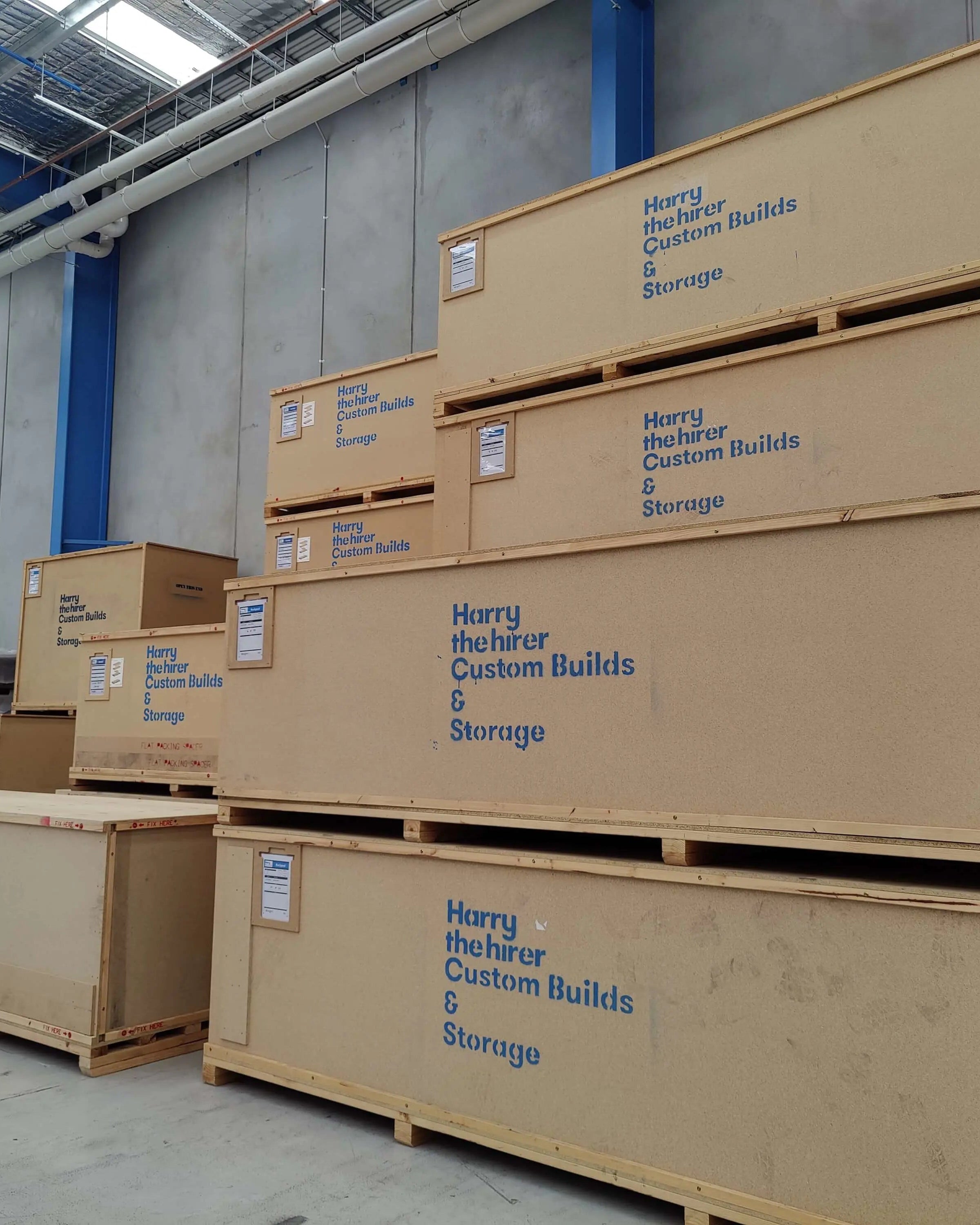 Stack of beige storage containers with 'Harry the Hirer Custom Builds & Storage' branding in a warehouse.