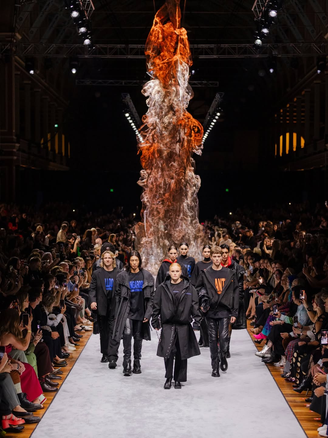 Fashion show with models walking down a runway under a large fiery sculpture.