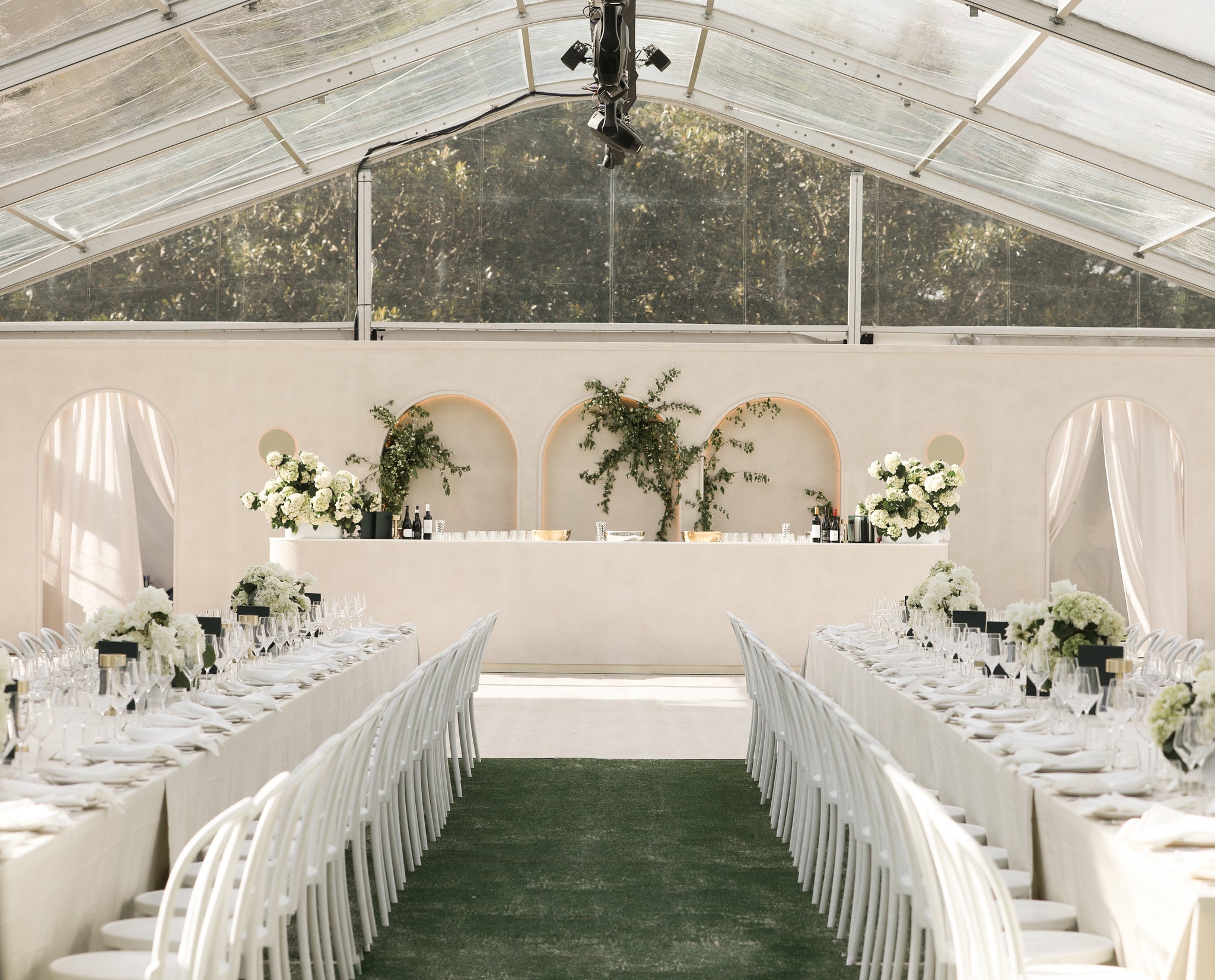 Wedding reception setup with tables, chairs, and floral arrangements inside a clear marquee