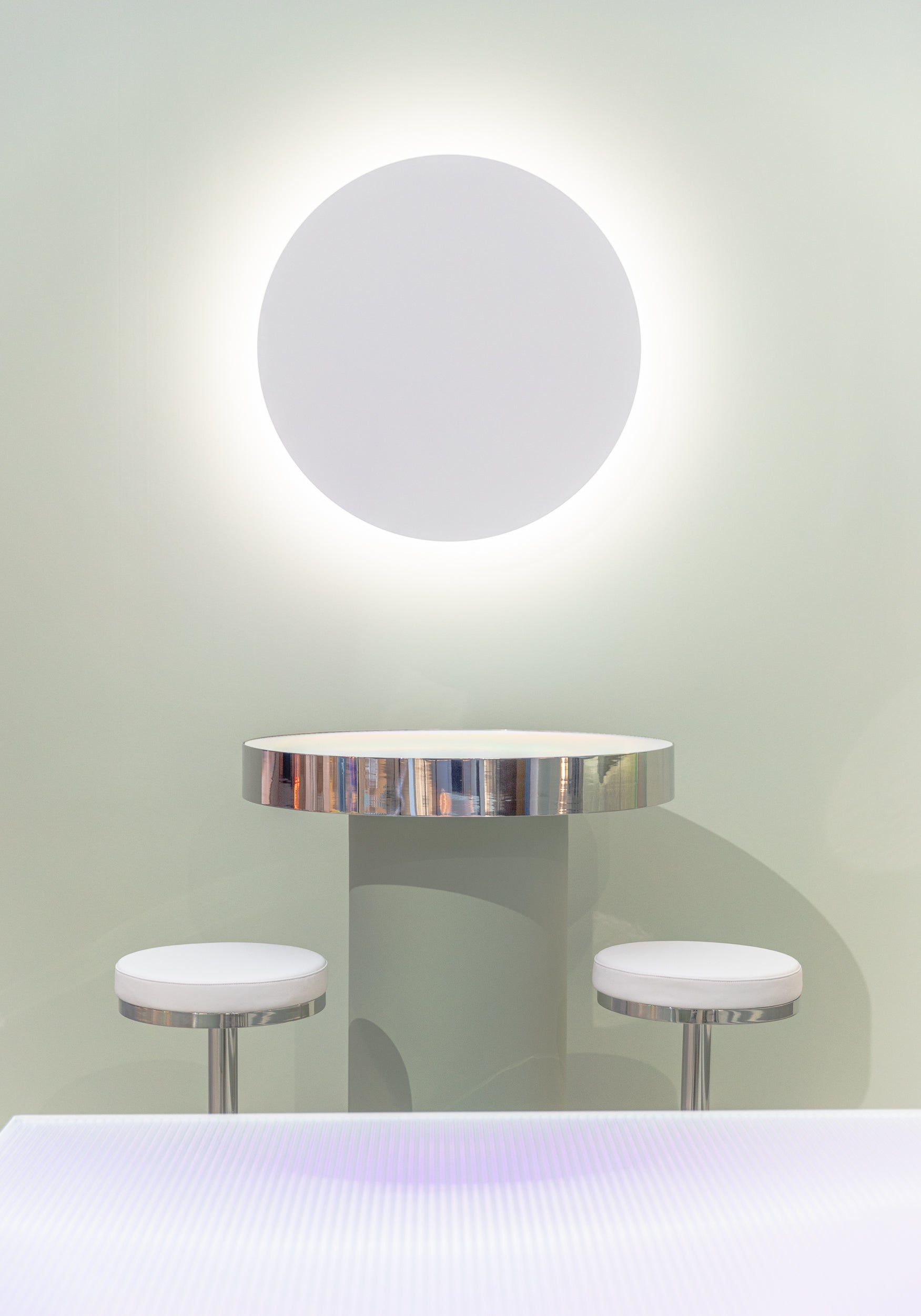 Round table with chrome rim and two stools against a light green wall with a circular light fixture.