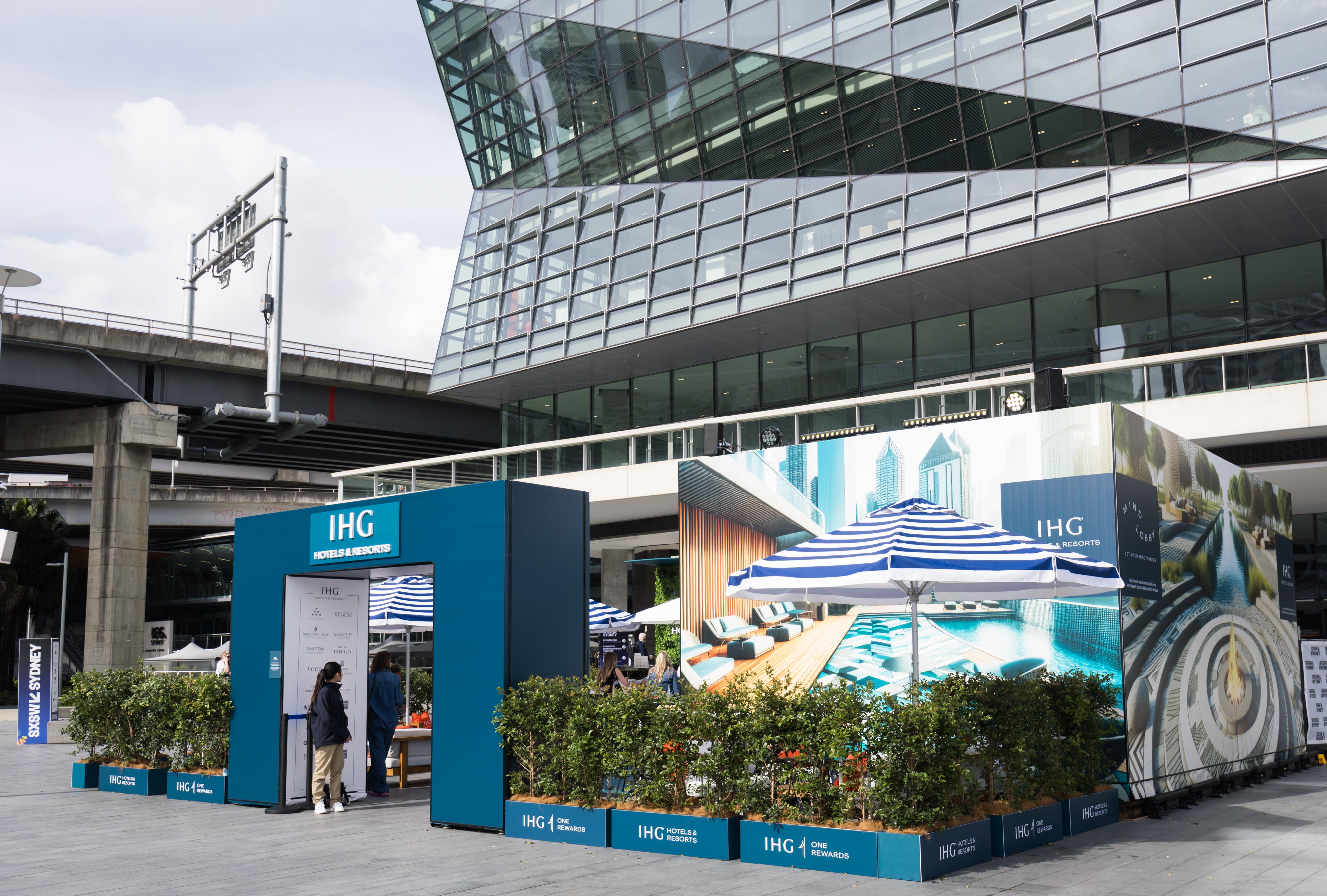 IHG promotional stand with large billboard featuring pool and beach scene in front of a modern building. All Harry The Hirer Signage