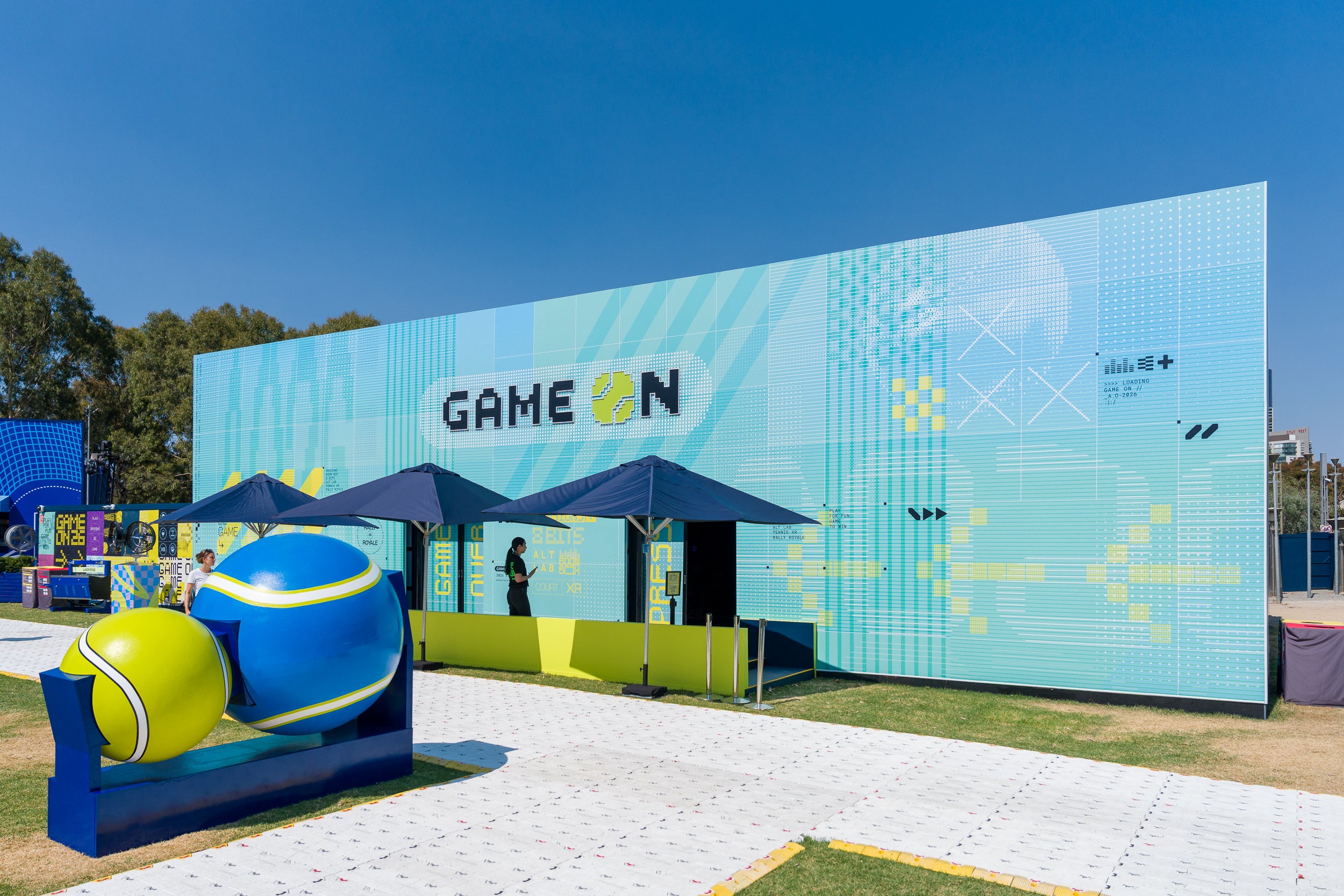 Outdoor event with 'GAME ON' branded backdrop and large inflatable tennis ball sculpture.