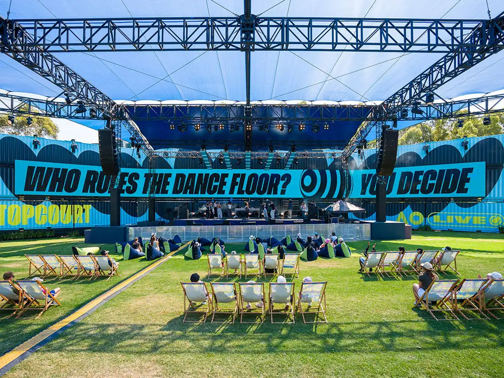 Outdoor event with 'WHO RULES THE DANCE FLOOR?' banner and people sitting on chairs.