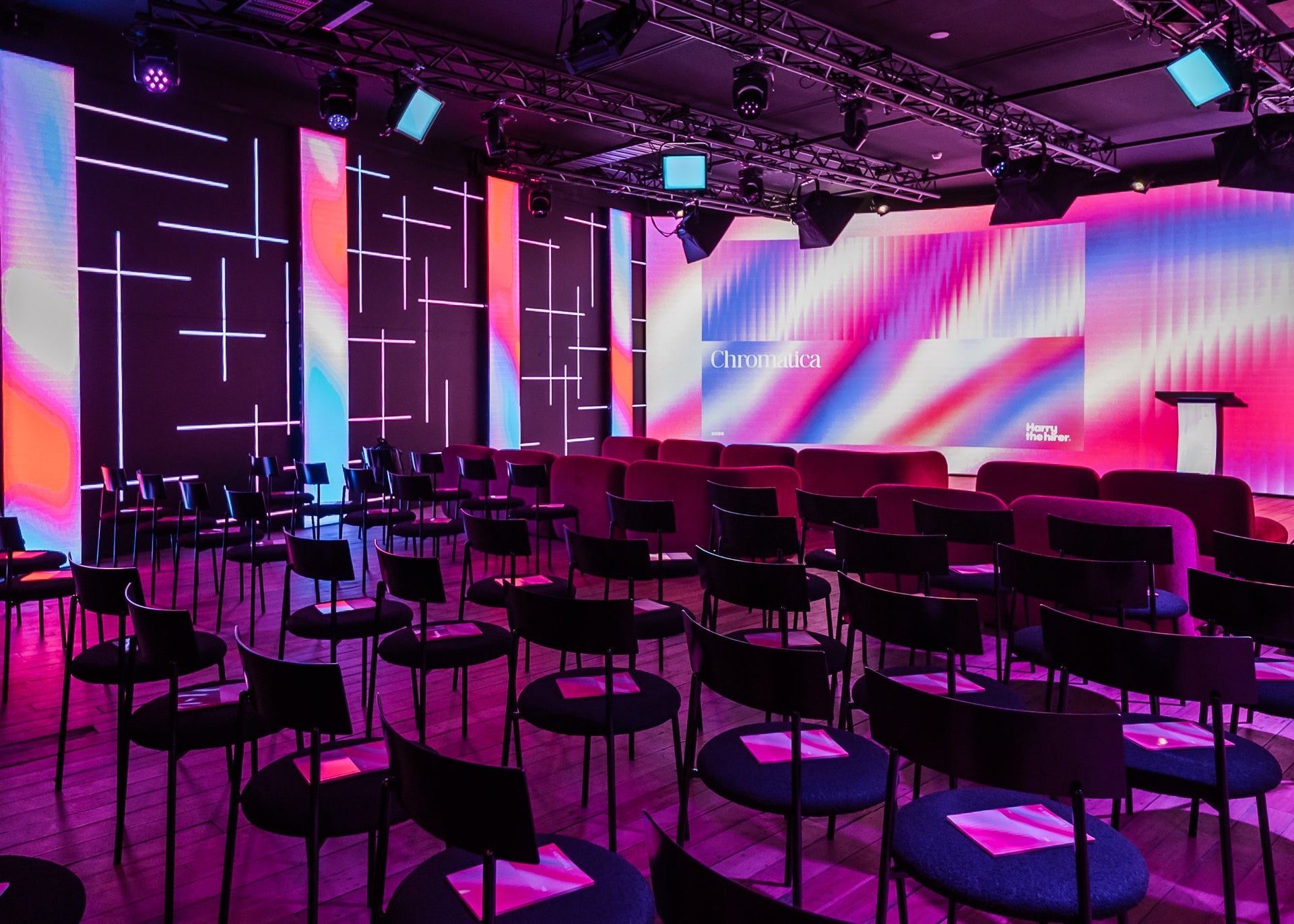 A modern event stage or auditorium interior with rows of black chairs and small round tables facing a large illuminated screen. The space is lit with vivid pink, purple, blue, and orange lighting, and the walls feature vertical LED panels with geometric line patterns.