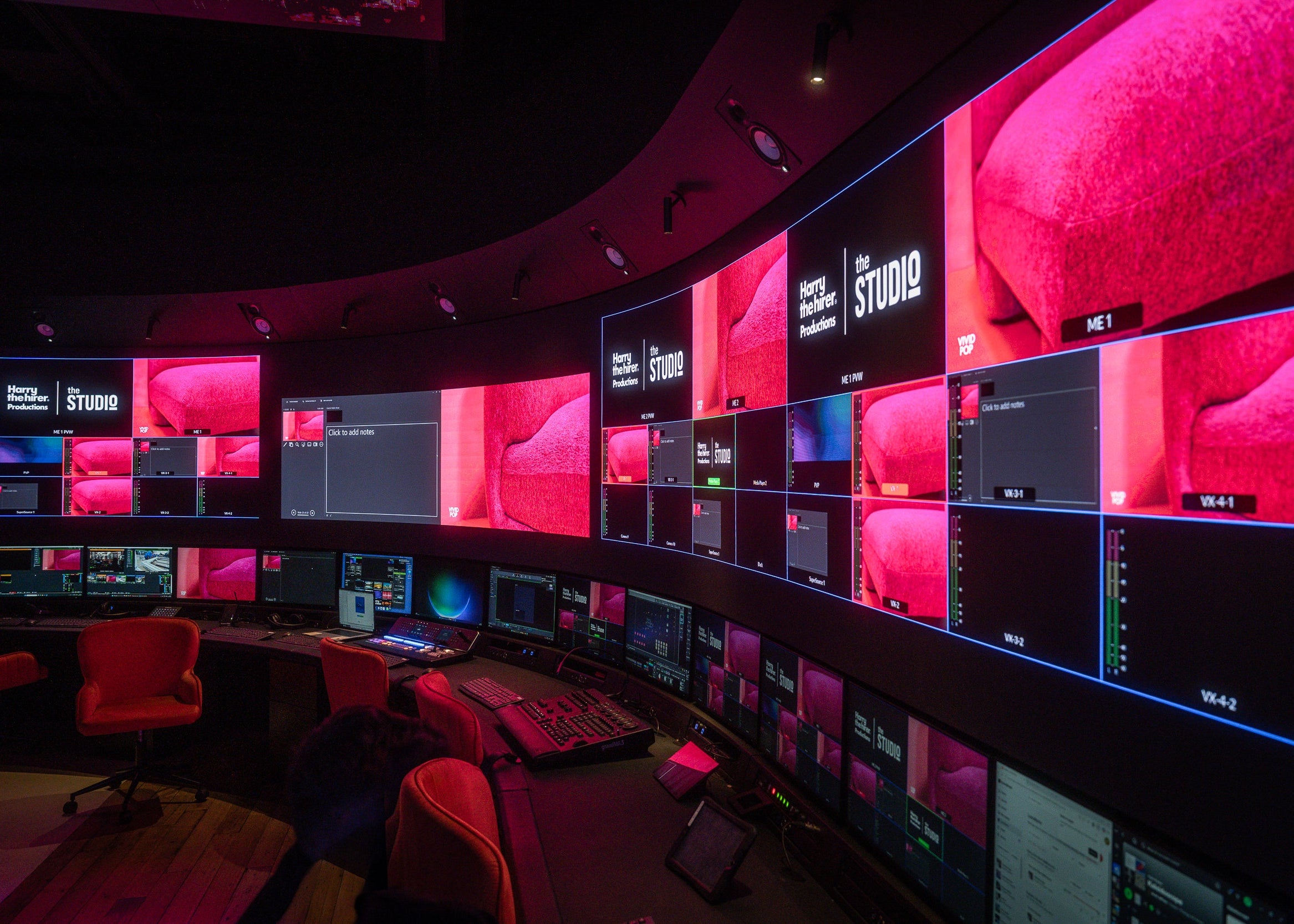 A dark control room with multiple curved video walls, monitors, mixing consoles, and red chairs in a studio environment.