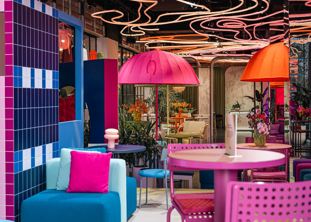 A vibrant, colorful modern showroom interior with bright pink umbrellas, pink and blue tables, a blue armchair with a hot pink pillow, and a decorative tiled wall.