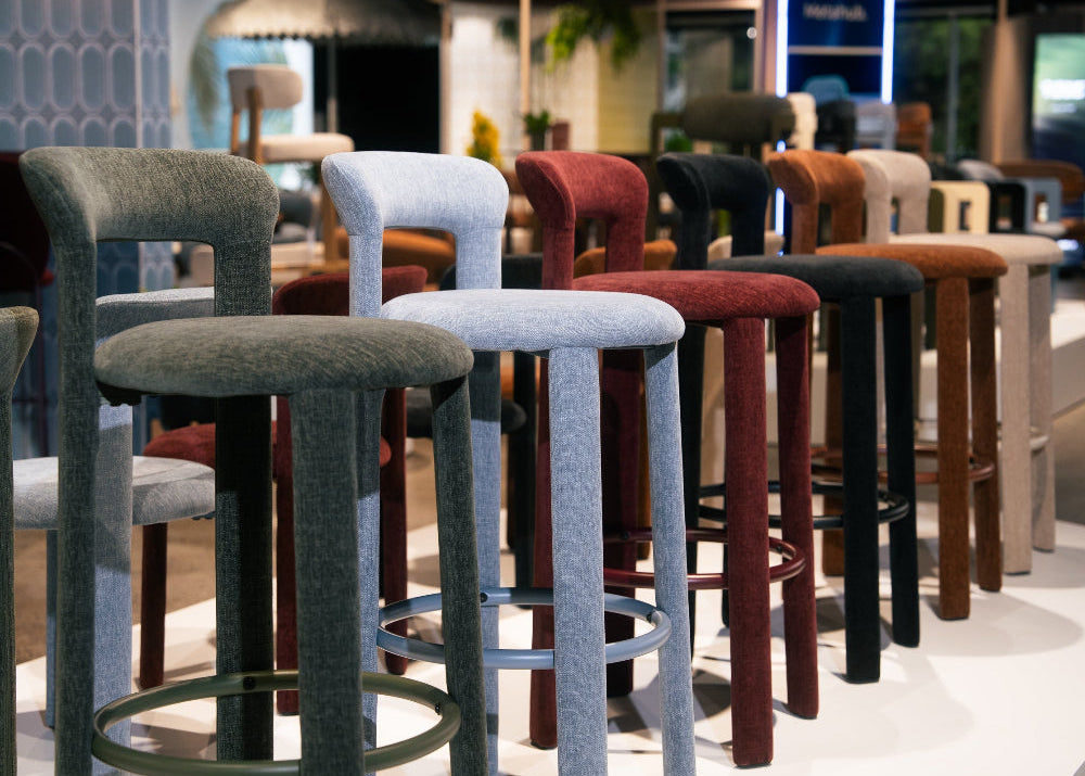 A row of modern bar stools in multiple colors, including gray, light blue, burgundy, black, brown, and beige, displayed on a white showroom floor.