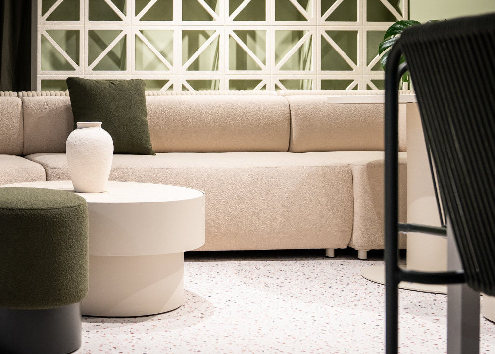 A modern beige sectional sofa in a styled showroom setting, featuring a dark green accent pillow, a matching green cylindrical ottoman, a white round coffee table, and a green outdoor rope chair in the foreground.