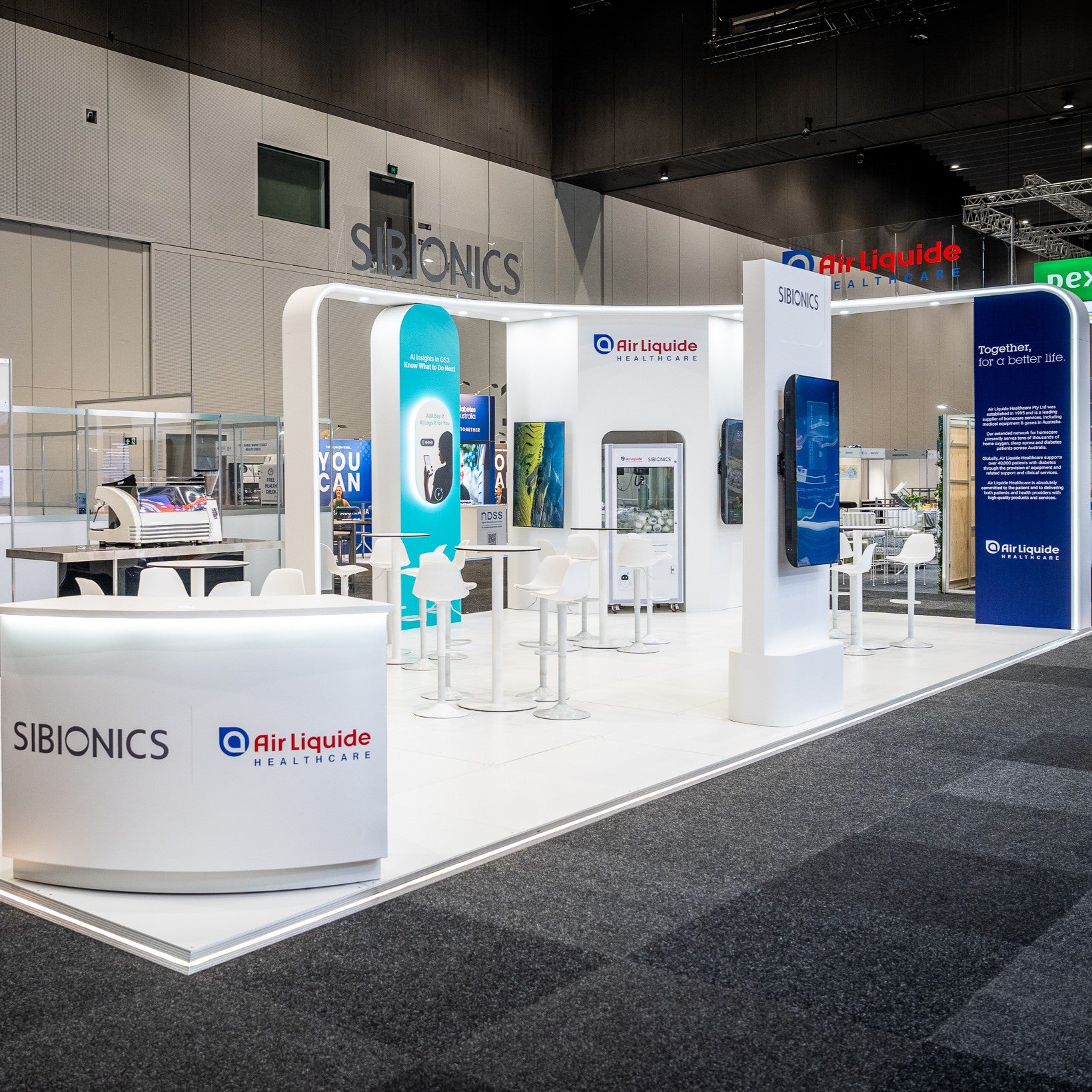 A large exhibition booth display for Sibionics and Air Liquide Healthcare, featuring a white reception desk, white display columns, high-top tables, stools, and medical device mockups on a carpeted trade show floor.