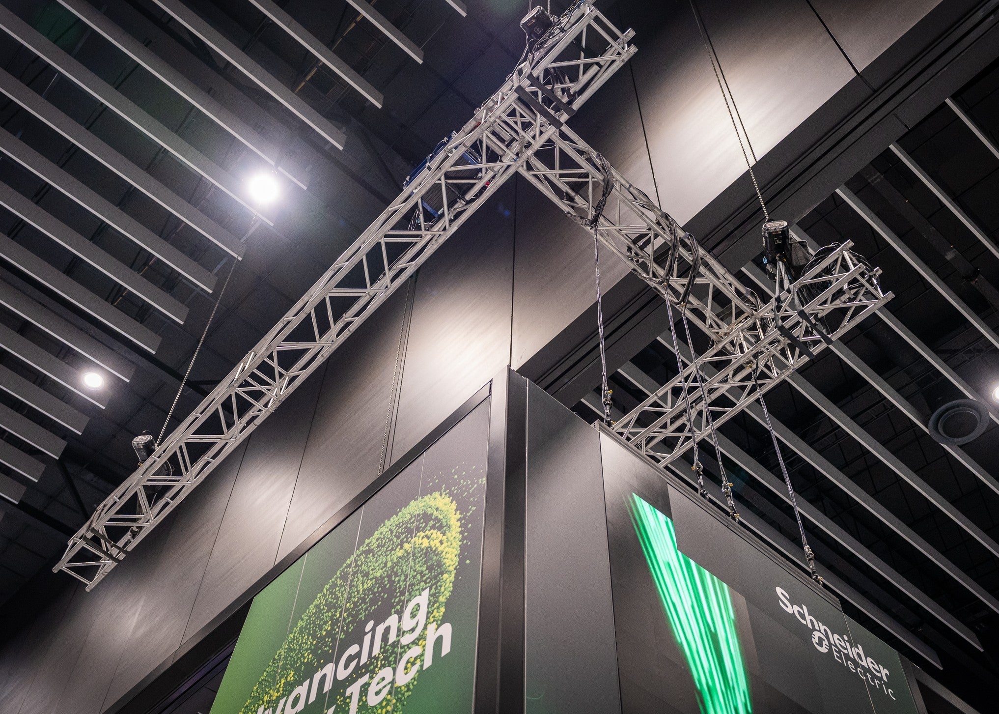 Large metal truss stage lighting rigs suspended from the ceiling of a convention center, with exhibition banners below featuring green graphics and text.