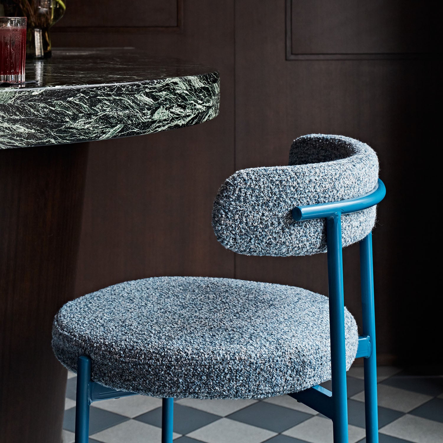Modern chair with blue frame and gray cushioned seat and backrest in a room with dark walls and a checkered floor.