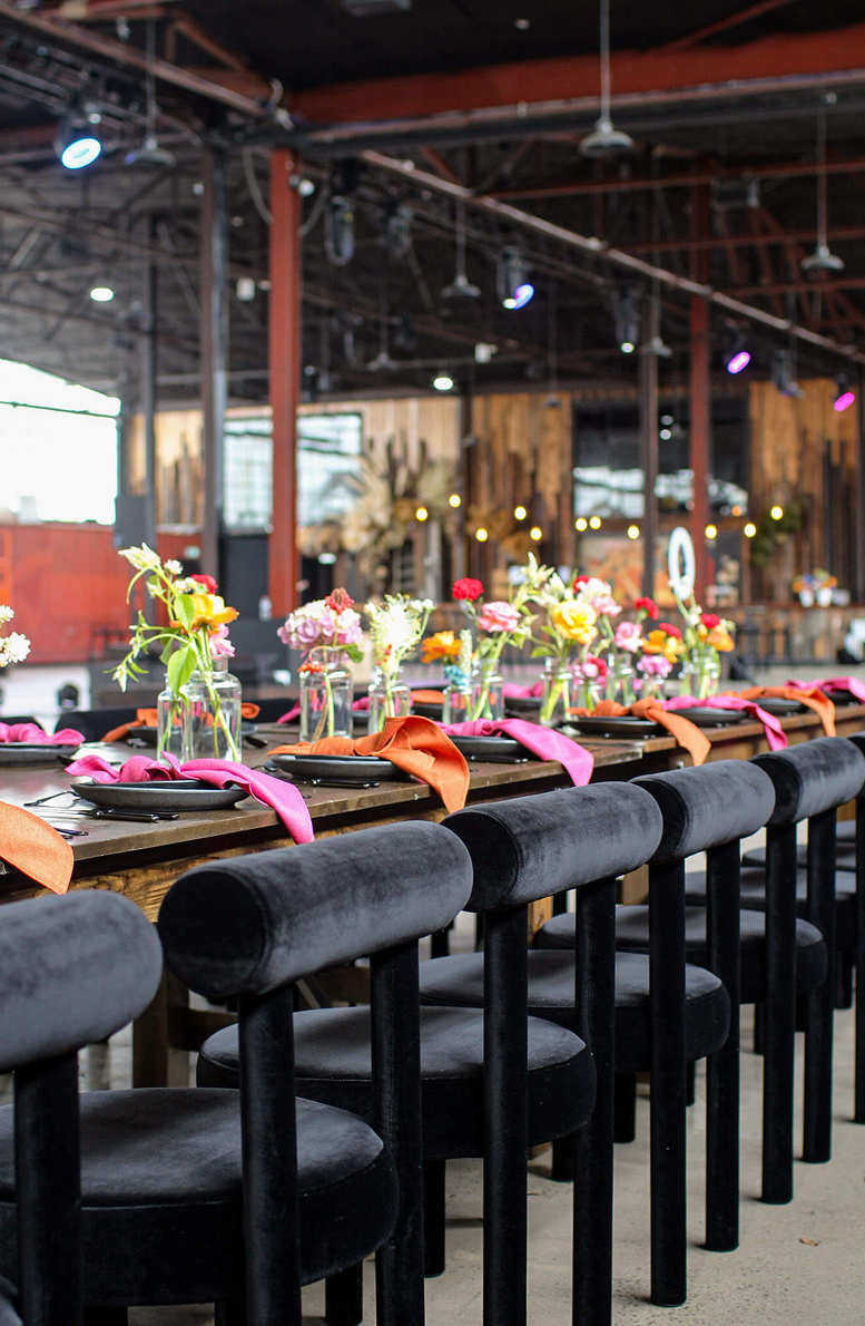 Long table set with floral arrangements and pink ribbons in a large industrial-style room.