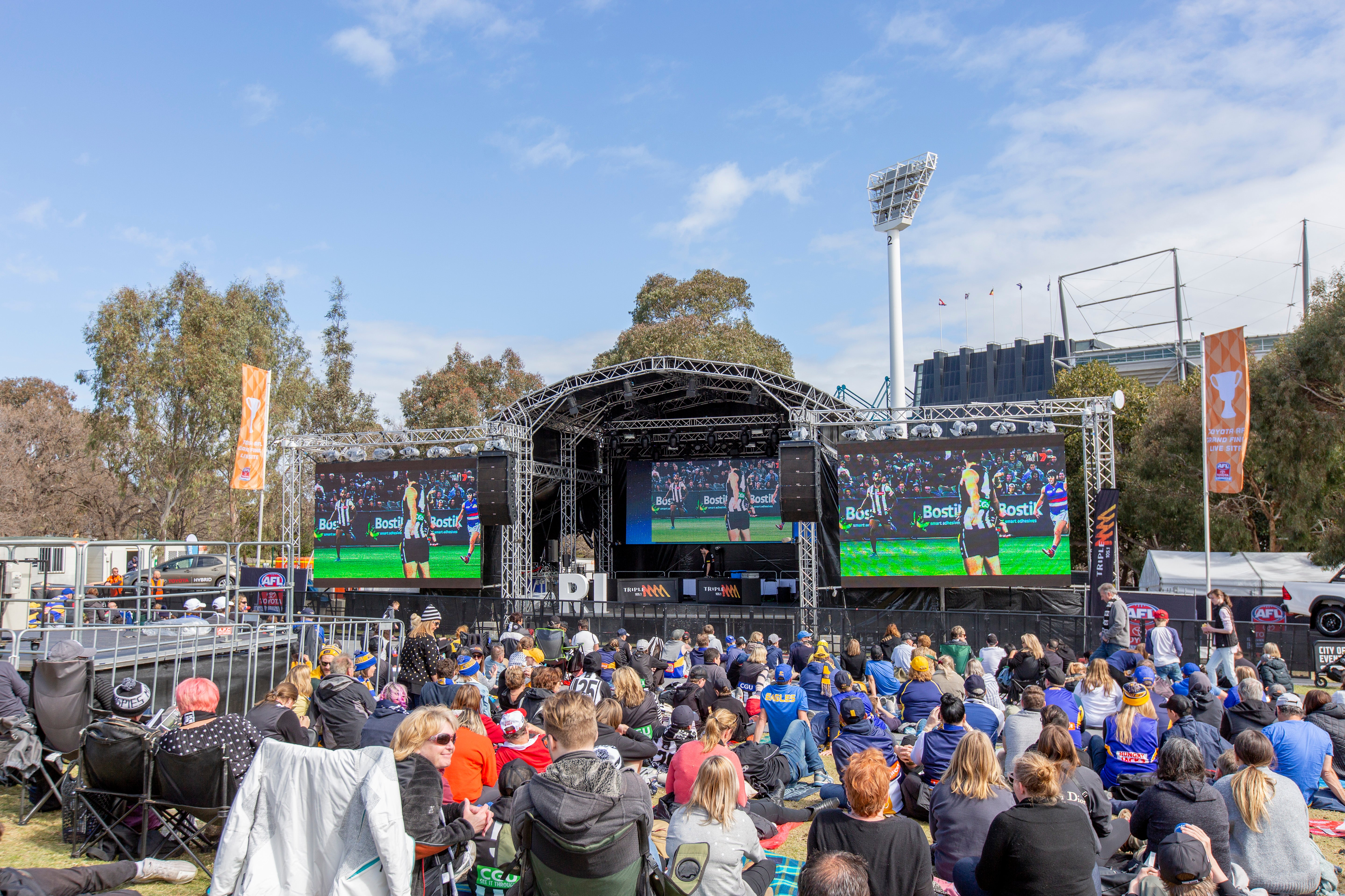 Outdoor event with a stage and large screens displaying the AFL grand final, surrounded by trees and clear sky.