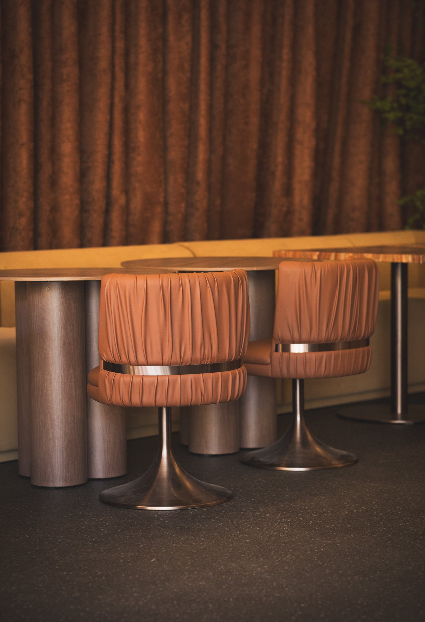 Two Rocco brown stools with metal bases in front of a wooden counter against a brown curtain.
