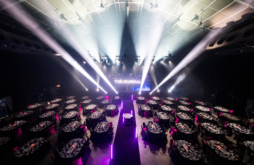 Dining setup with tables and chairs in a large hall, illuminated by spotlights.