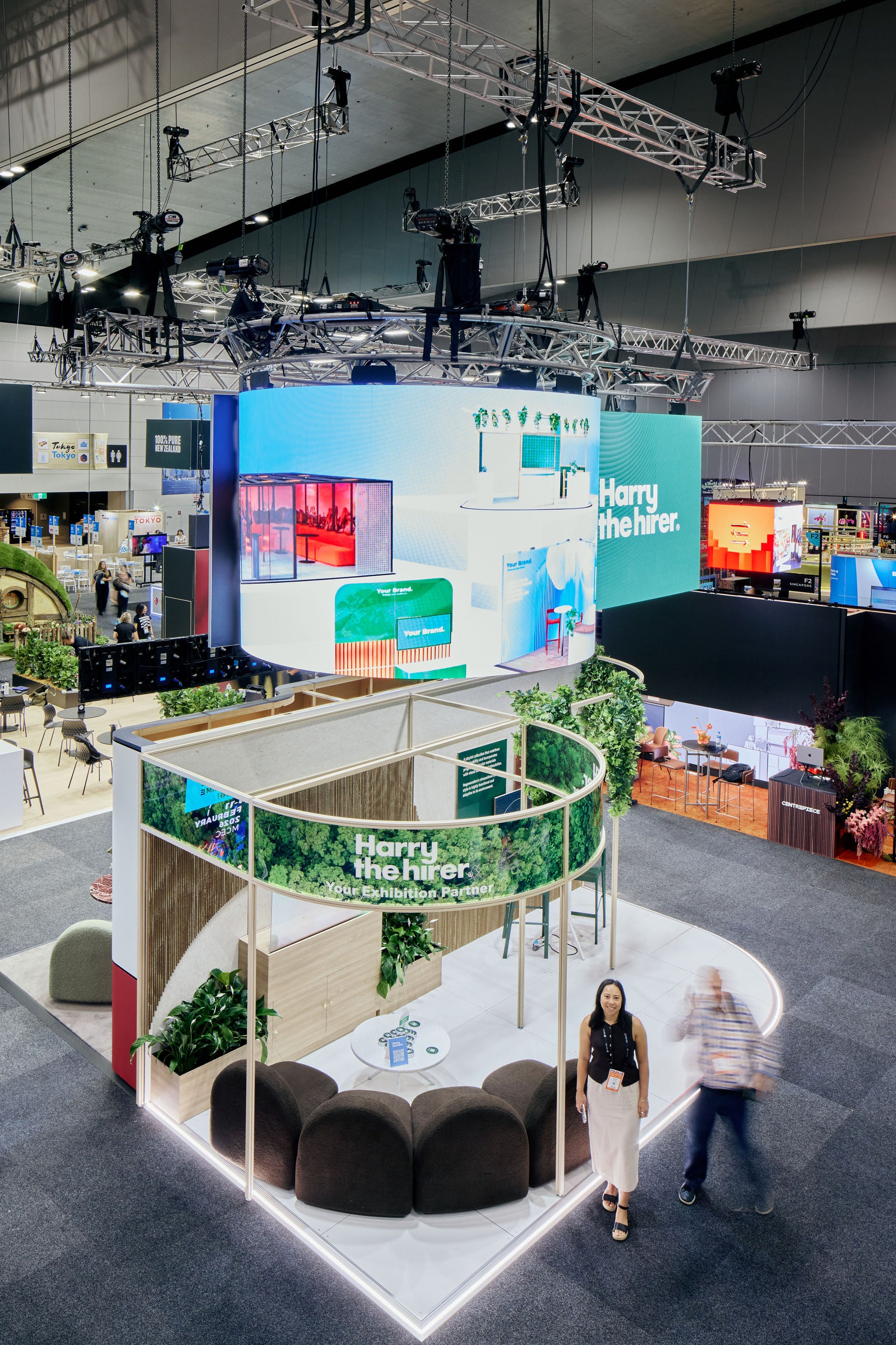 Modern indoor space with a large screen displaying 'Harry the Hirer' branding, people walking around, and various furniture.