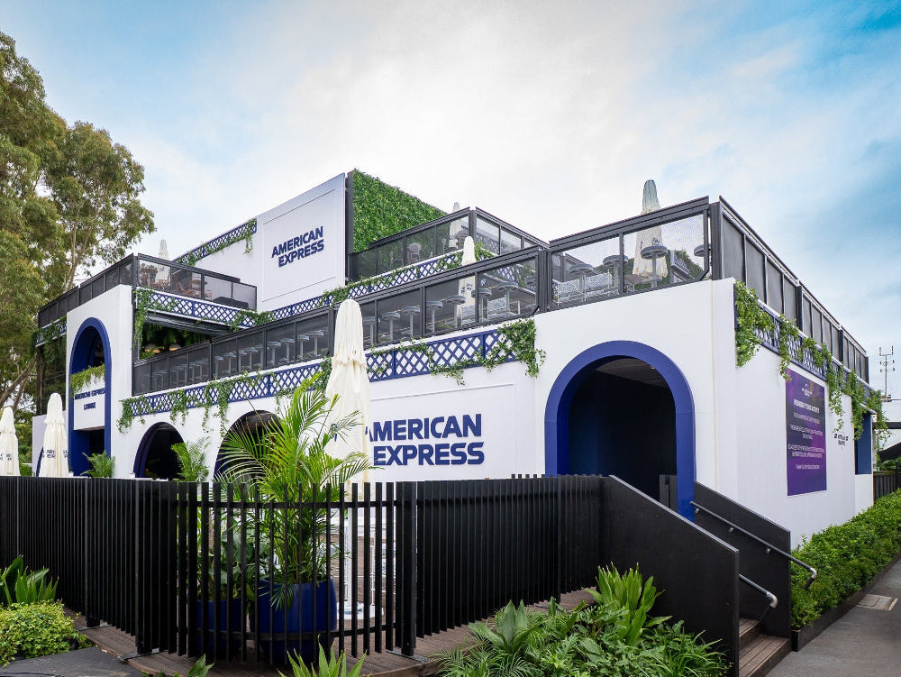 A two-story white and blue American Express building with outdoor seating, black railings, and surrounding greenery under a blue sky.