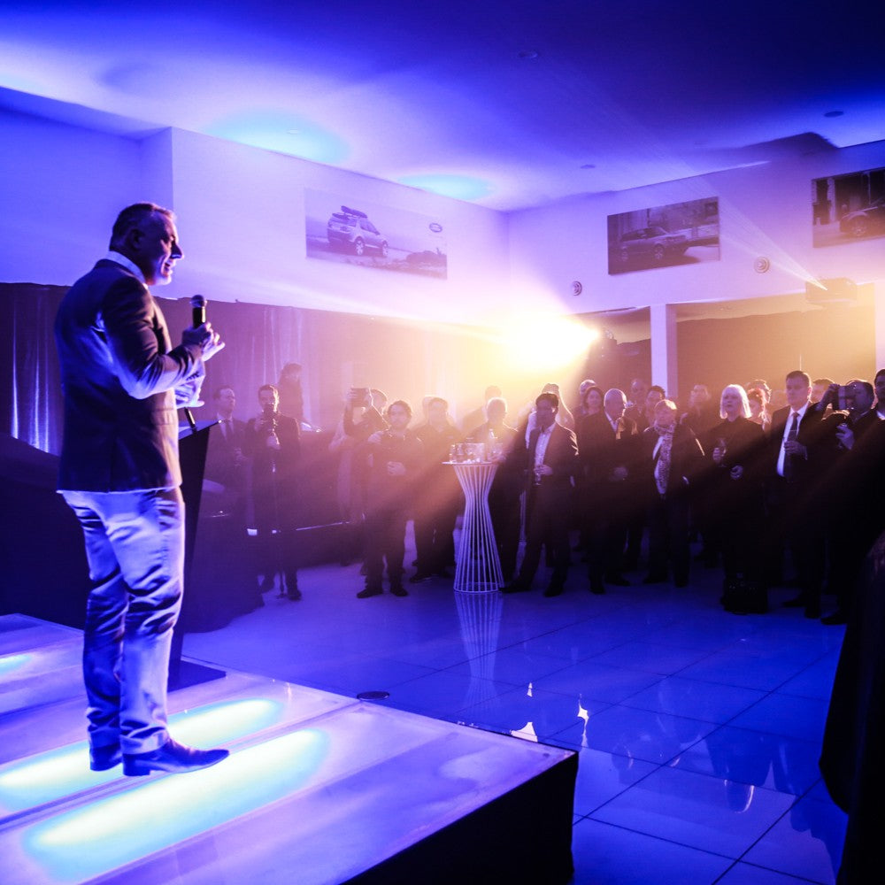 Man on a stage with a microphone addressing an audience in a dimly lit room.