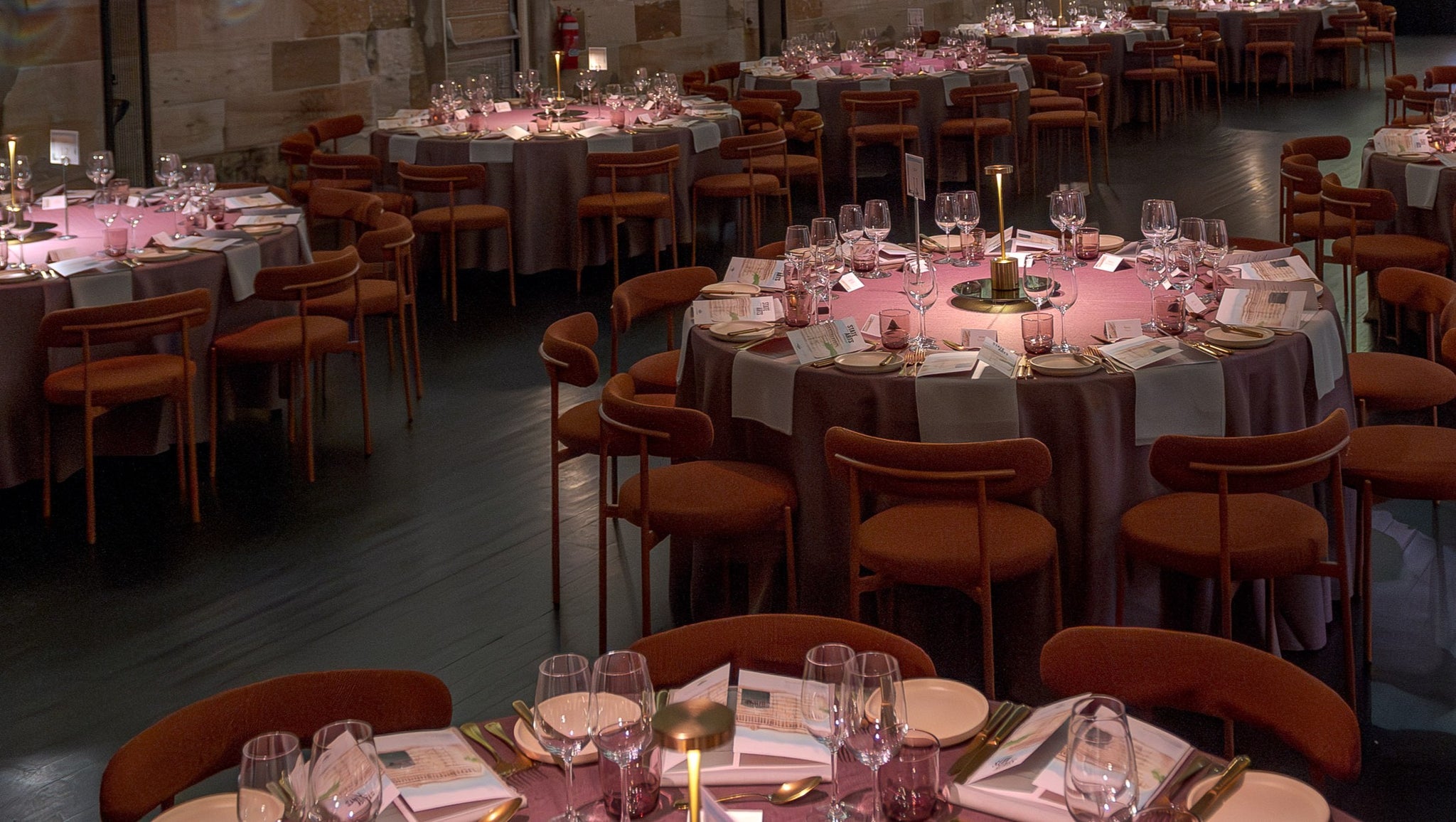 Dining hall with tables set for a formal event, featuring pink tablecloths and candles. Featuring our Margot chair, crockery and glassware.
