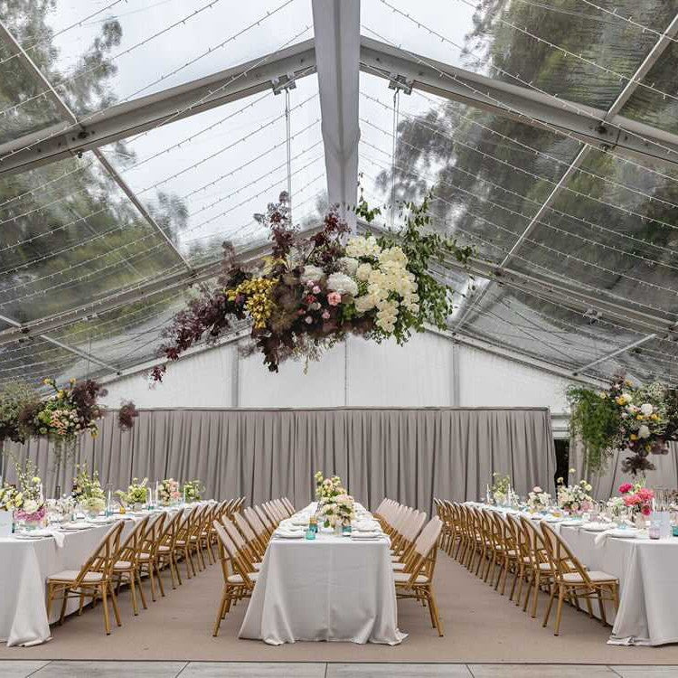 Decorated wedding reception with tables, chairs, and floral arrangements under a clear tent.