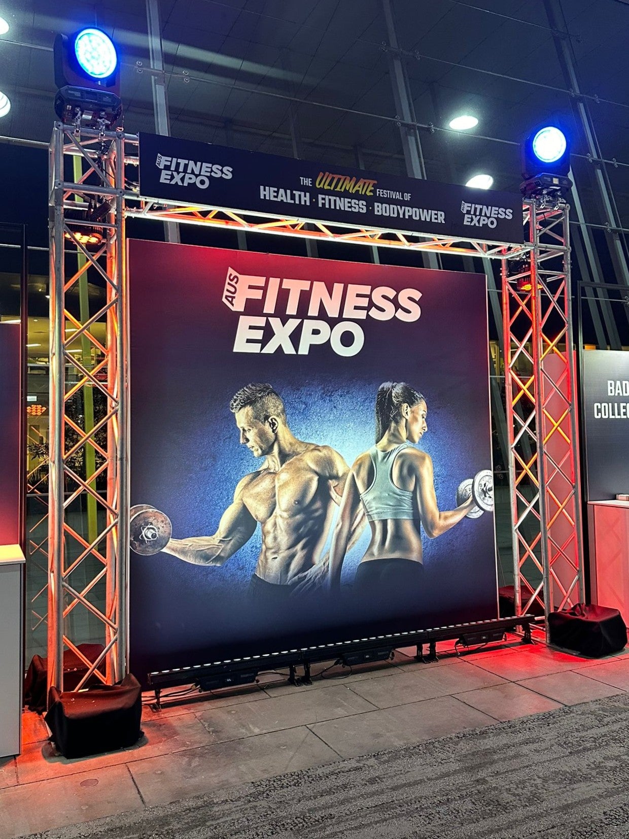 Fitness Expo banner with two muscular individuals lifting weights, surrounded by event setup.