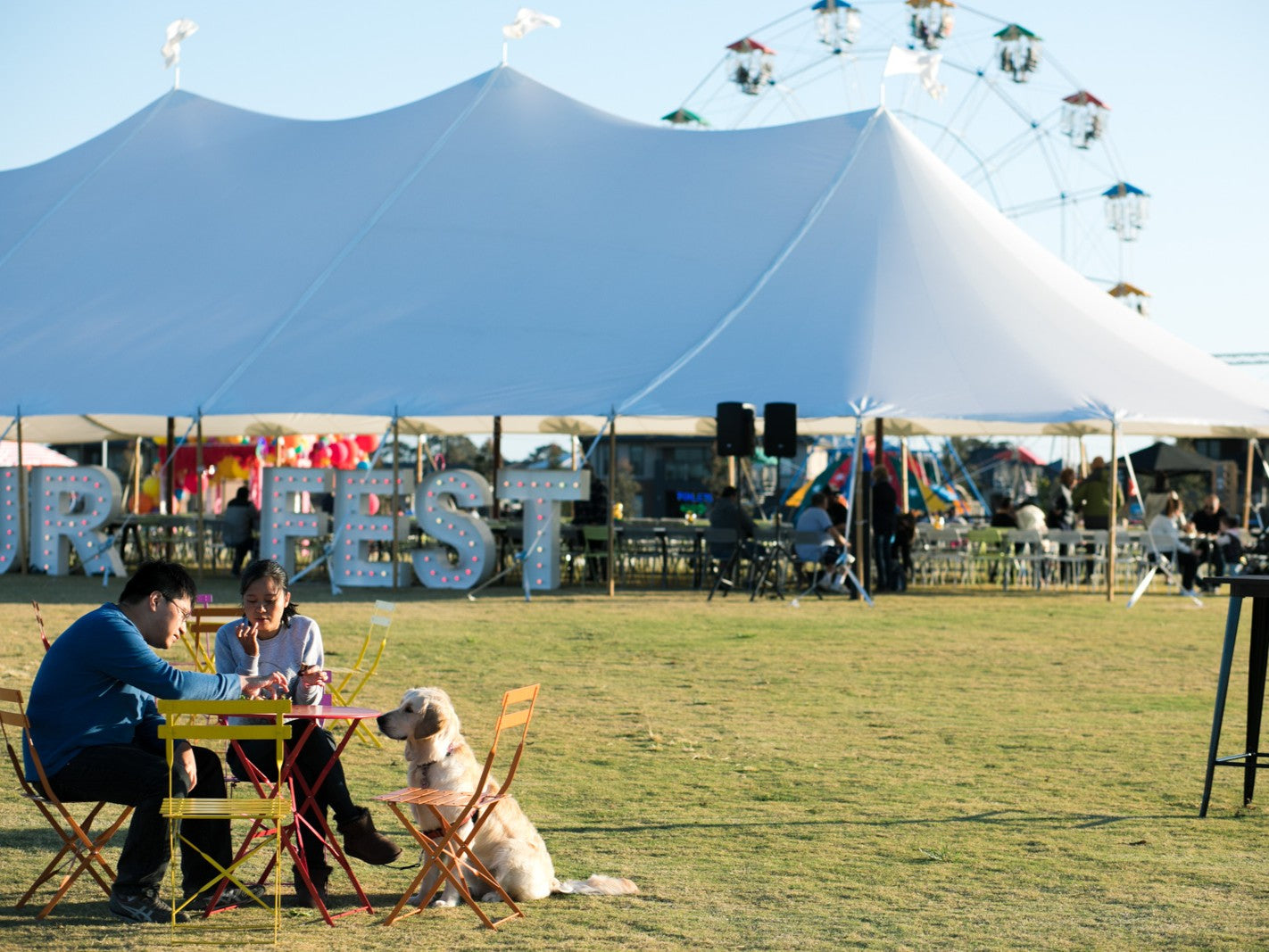 Two people sitting at a table with a dog on grass, with a Fairhaven Marquee in the background
