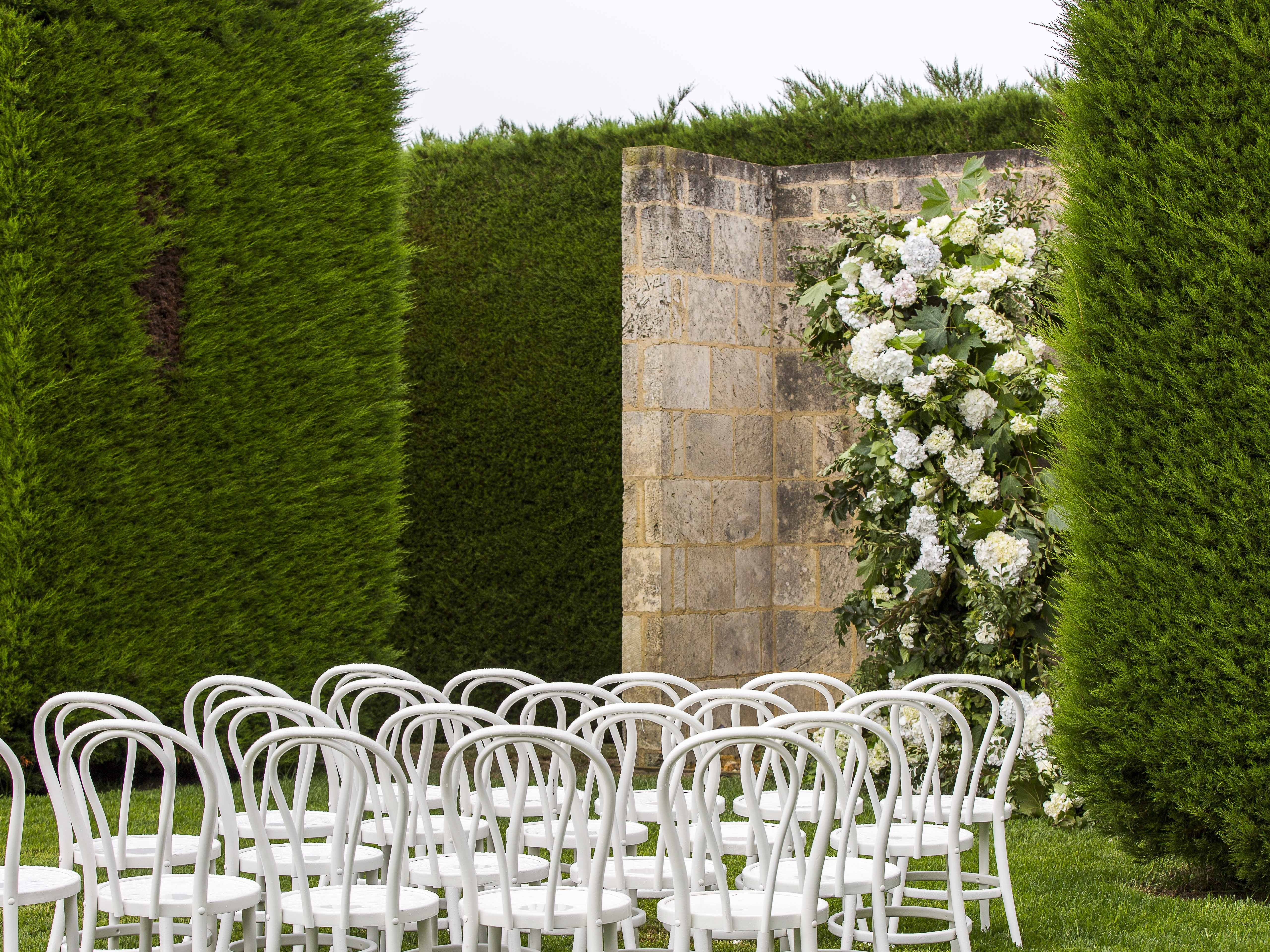 White chairs arranged for a wedding ceremony with floral decorations against a stone wall and greenery.