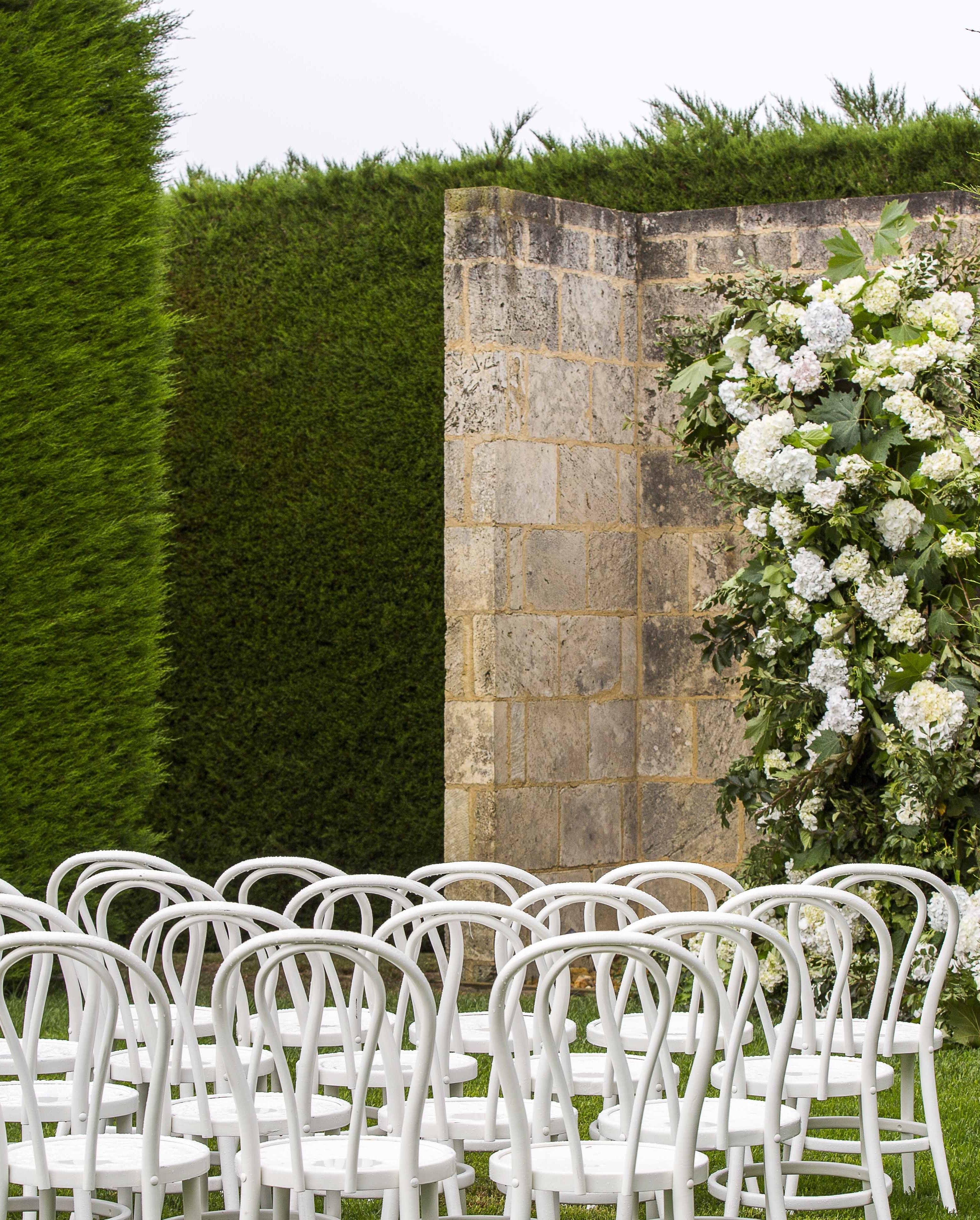 White chairs arranged for an outdoor ceremony with a stone wall and floral archway.