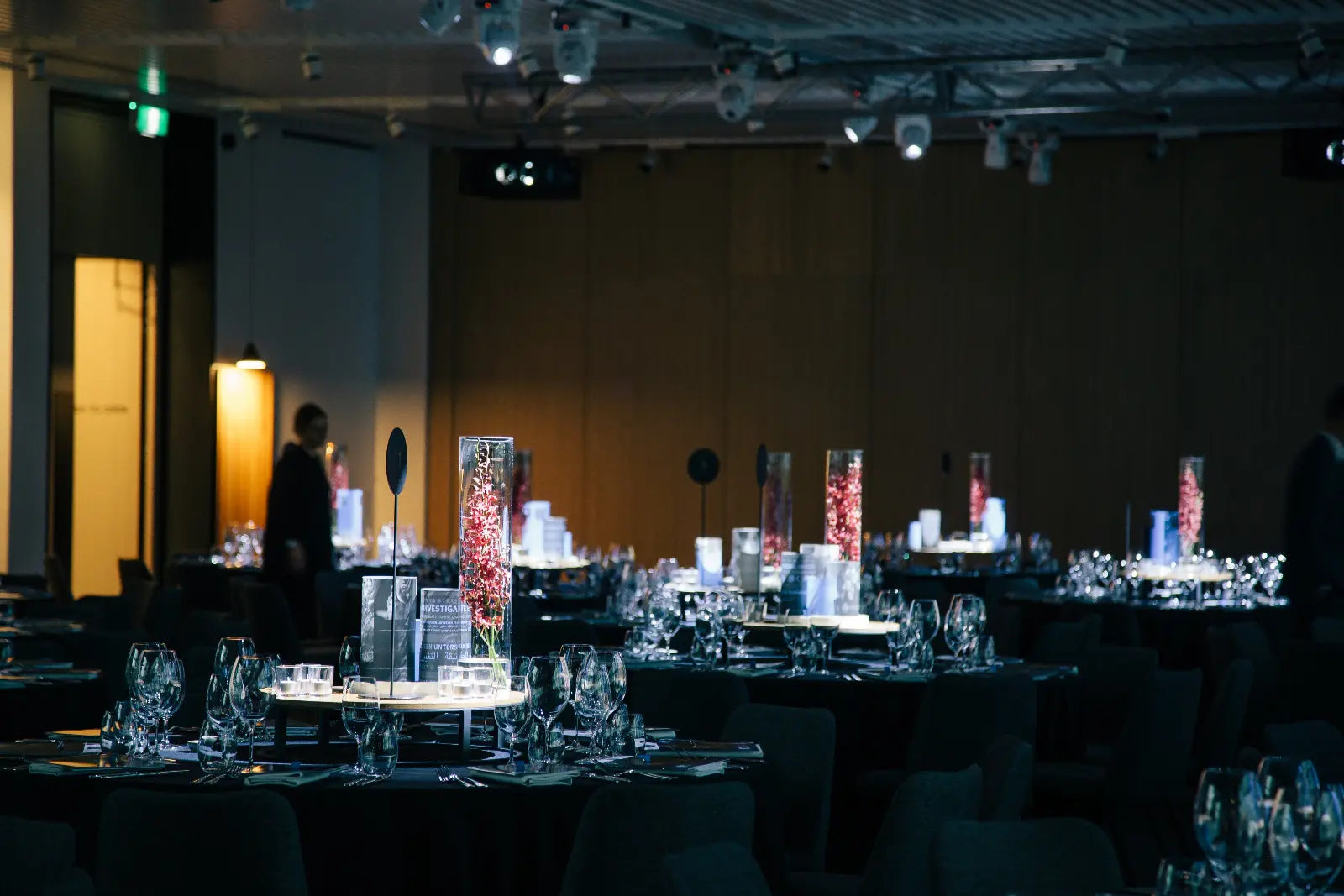 Dining room with tables set for a formal event, featuring tall glass vases with decorative elements.