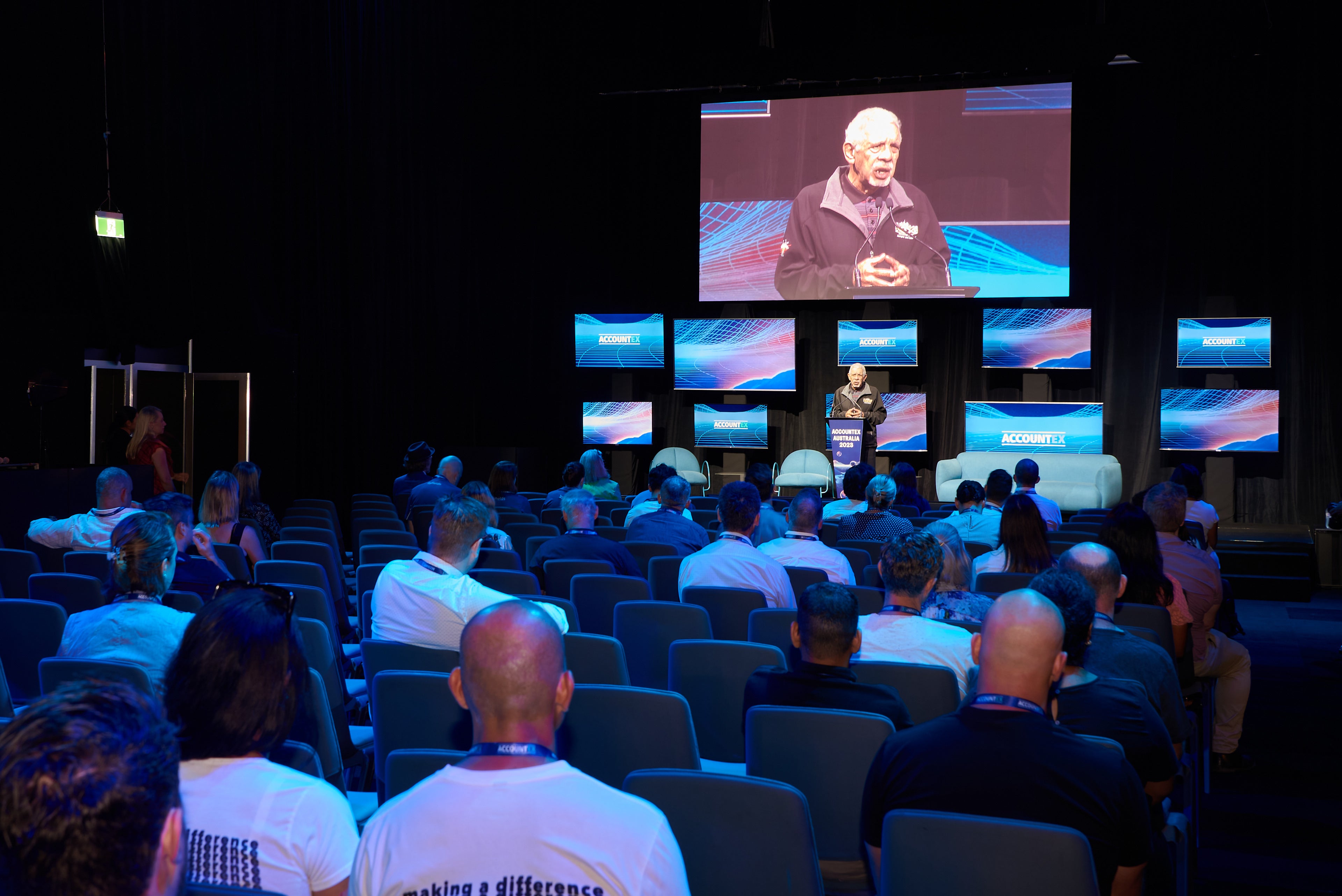 Audience in a conference hall watching a speaker on a large screen.